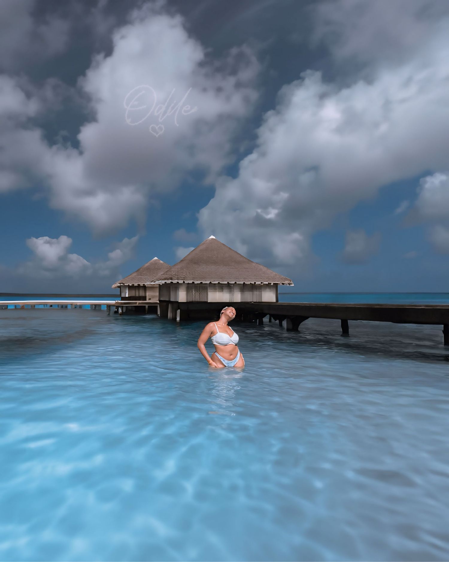 A figure in white swimwear poses in turquoise waters near overwater bungalows at a tropical resort under cloudy skies.