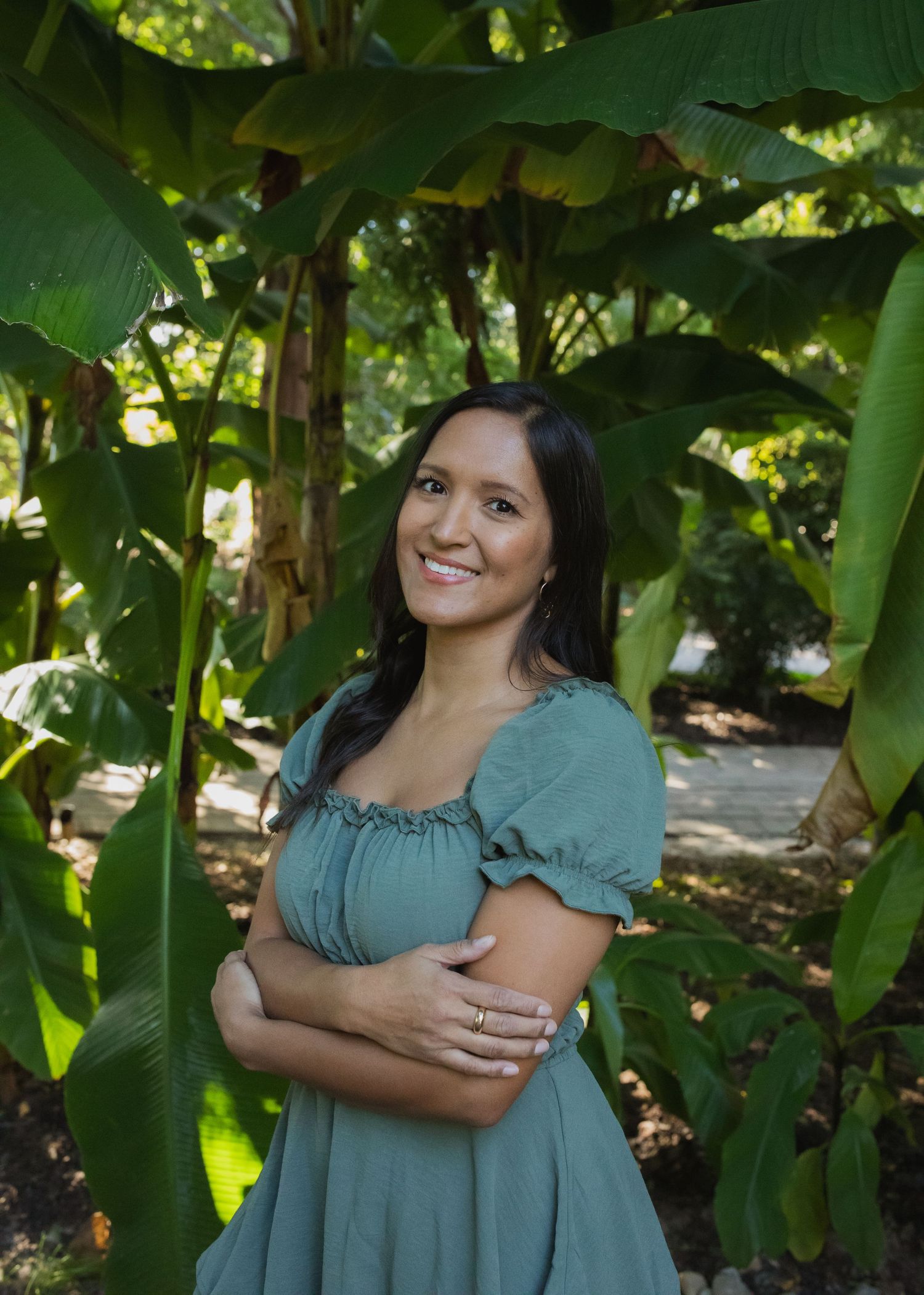 Person in a teal dress poses among tropical banana leaves in a garden setting.