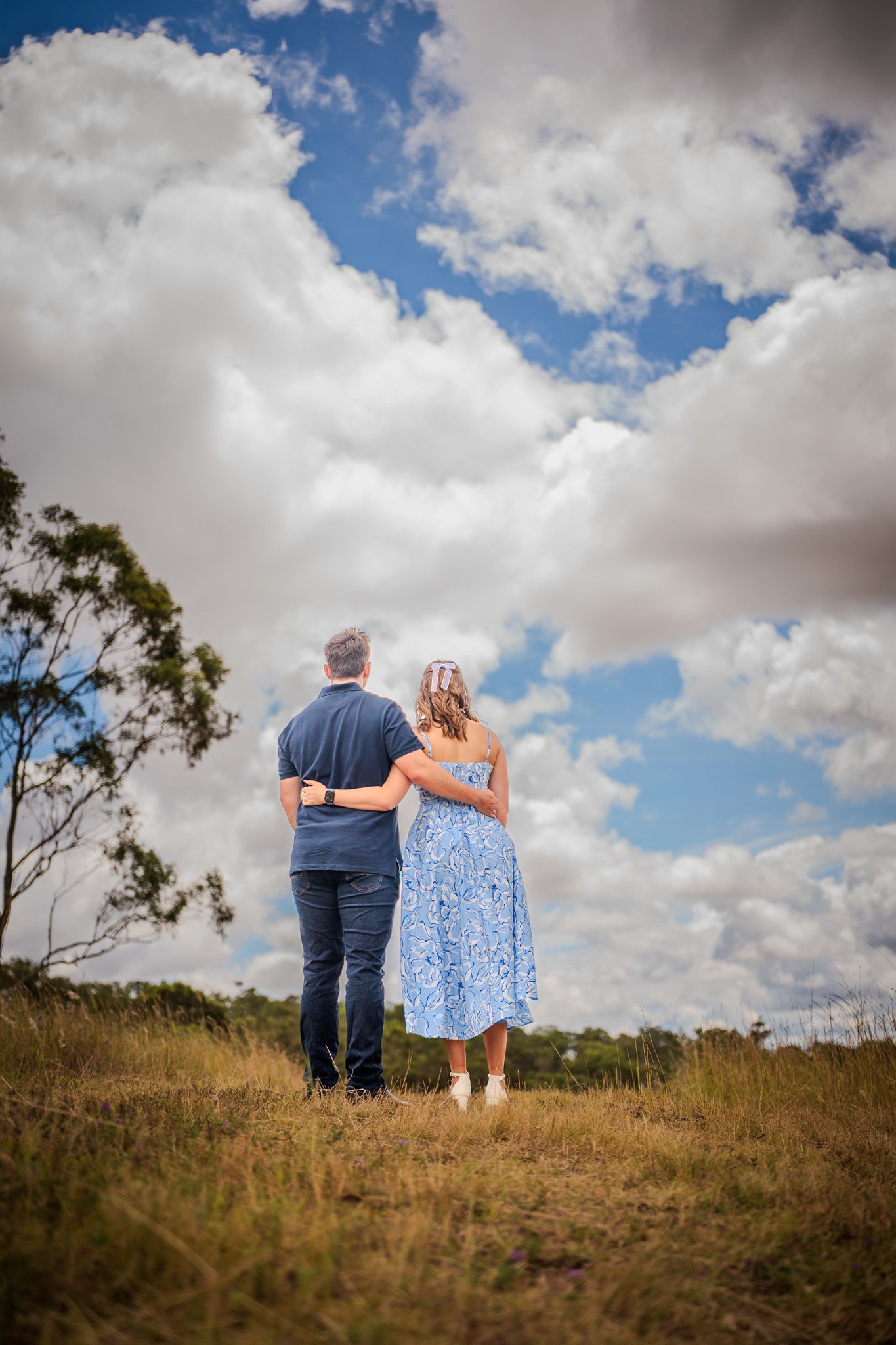 A couple stands together on a grassy hill overlooking scenic views under dramatic cloudy skies.