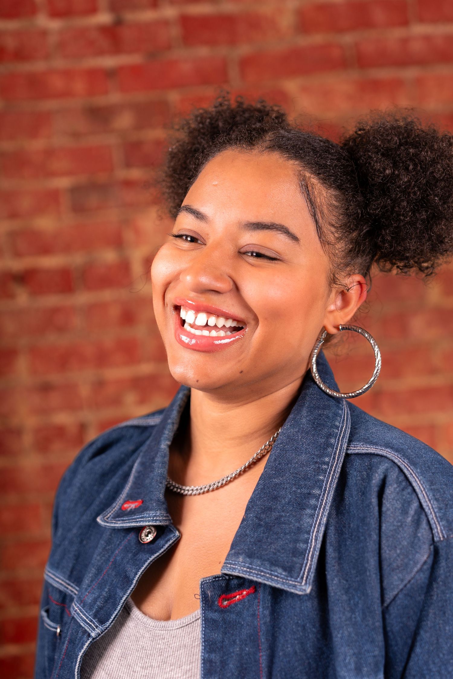 Person wearing denim jacket and silver hoop earrings smiles warmly against red brick wall background.