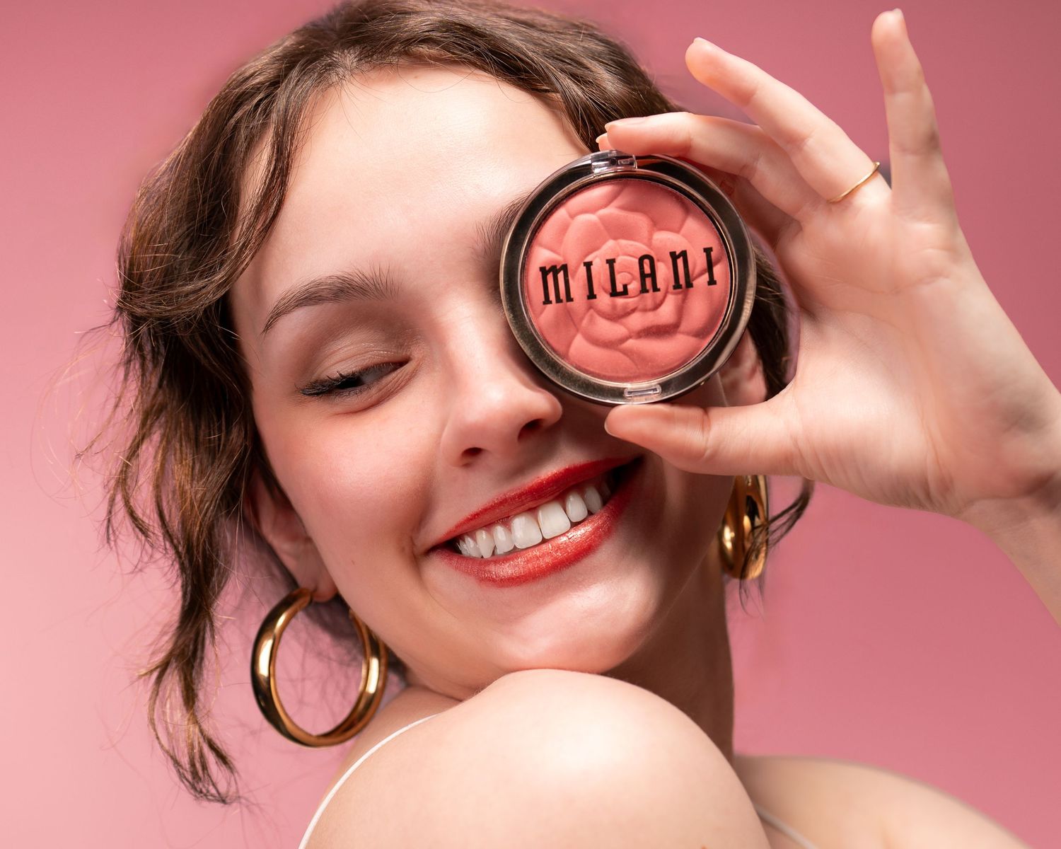 A beauty product photoshoot shows a person holding a Milani compact against a pink background.