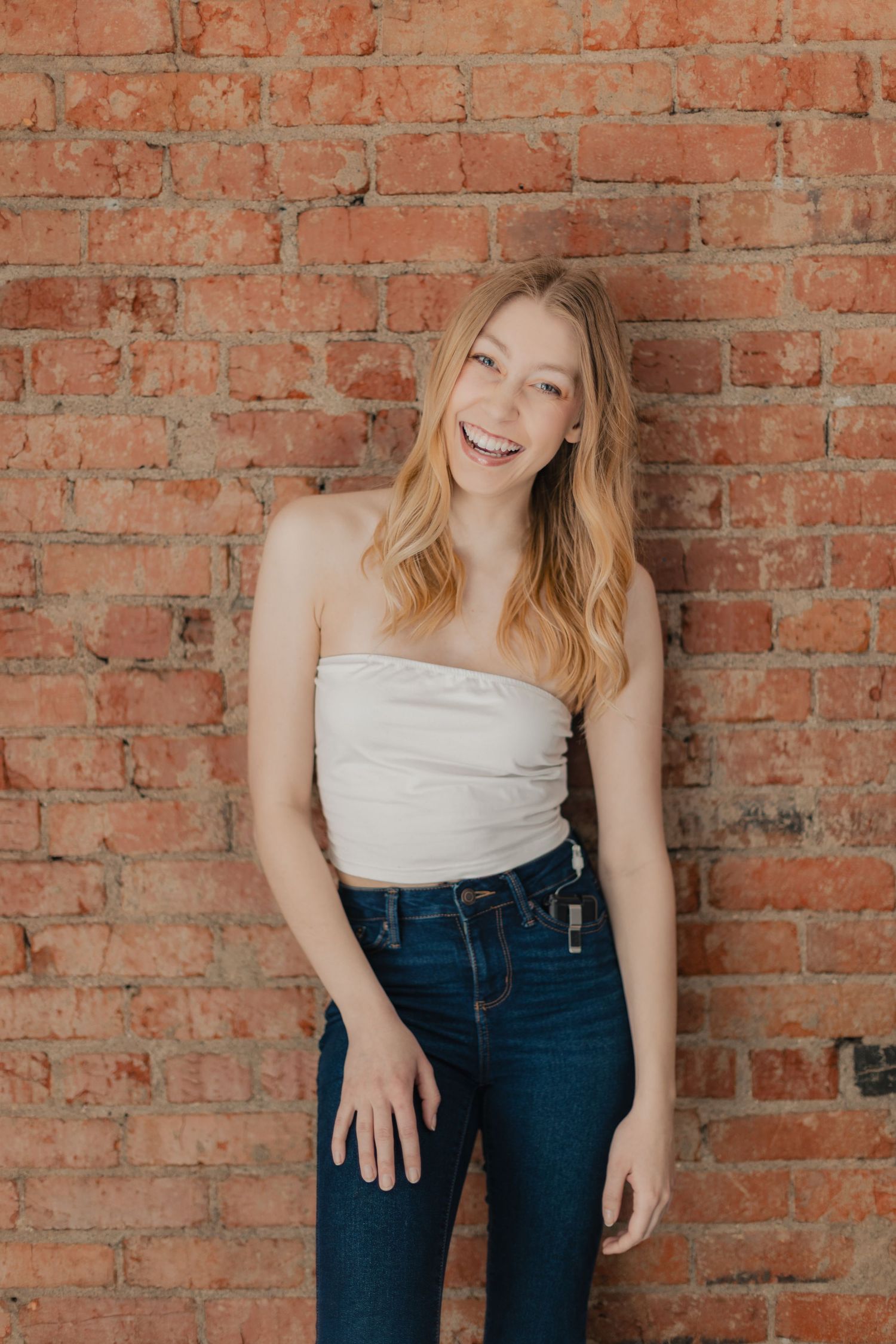 Young woman with long blonde hair smiling in white tube top and dark jeans against red brick wall.