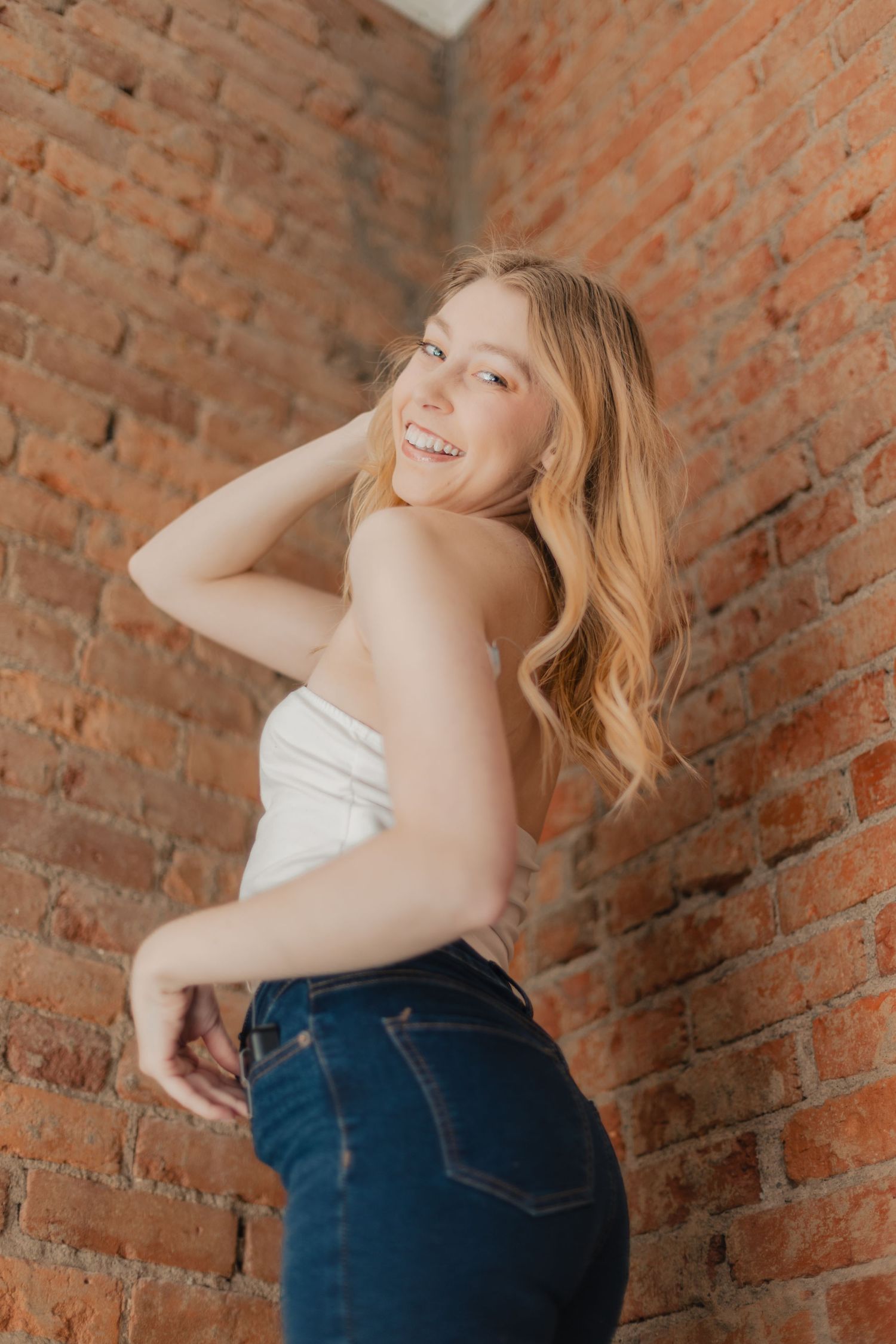 Smiling blonde woman posing in white tube top and dark jeans against rustic red brick wall.