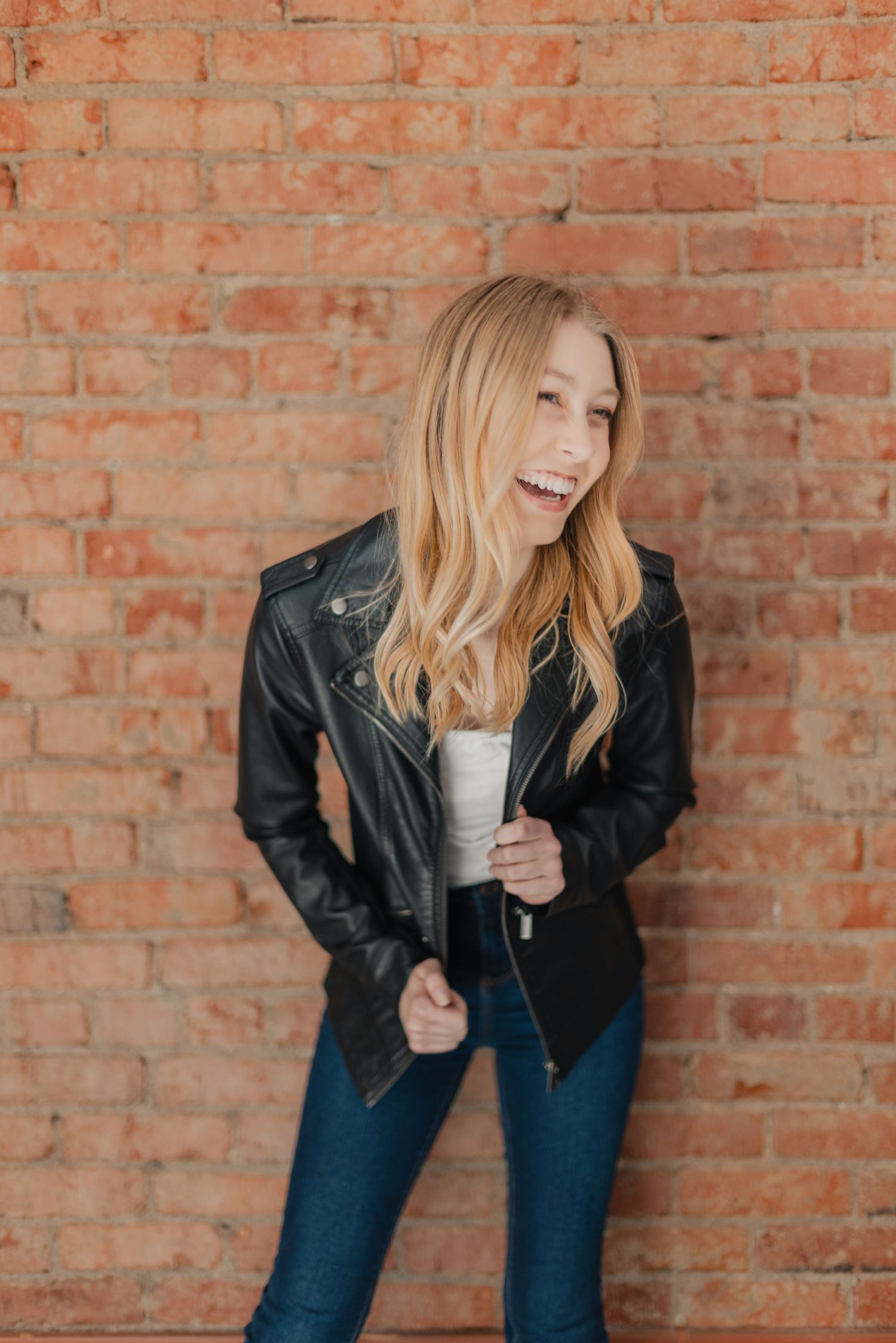 Laughing blonde woman in black leather jacket and jeans posing against a red brick wall.