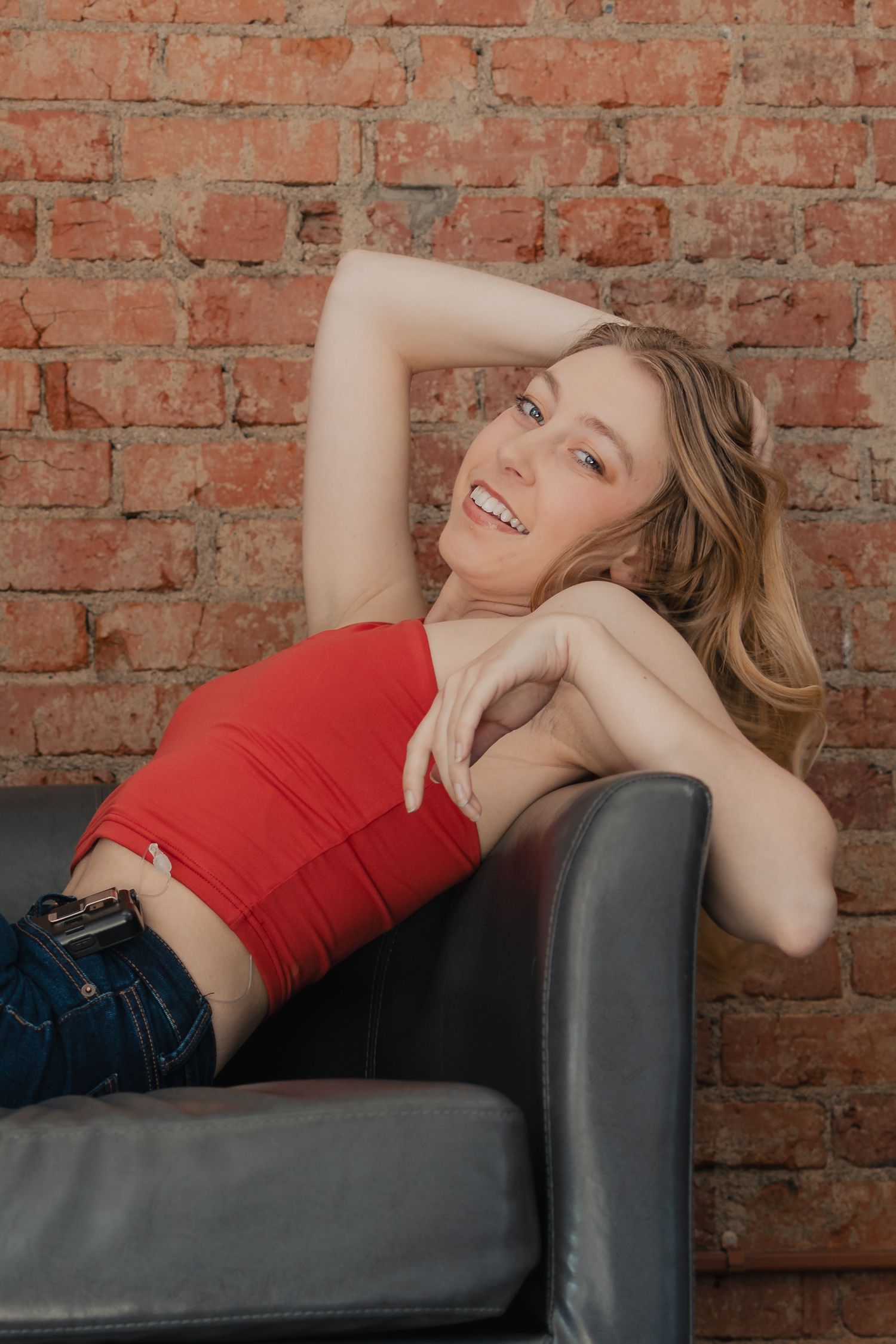 Young woman in red top and jeans relaxing on leather sofa against rustic brick wall, smiling cheerfully.