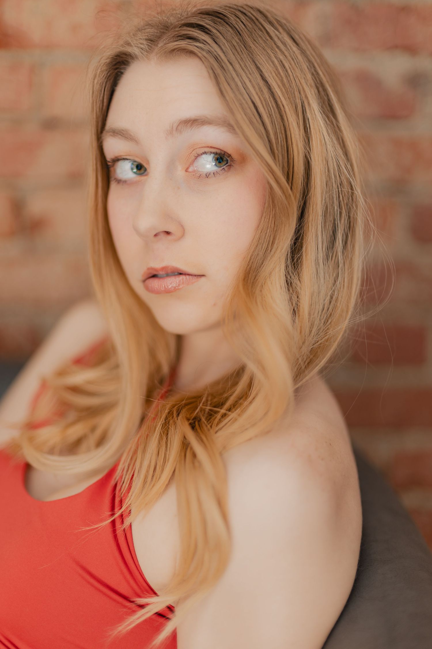 Young woman with blonde hair wearing a red top poses against a rustic brick wall background.