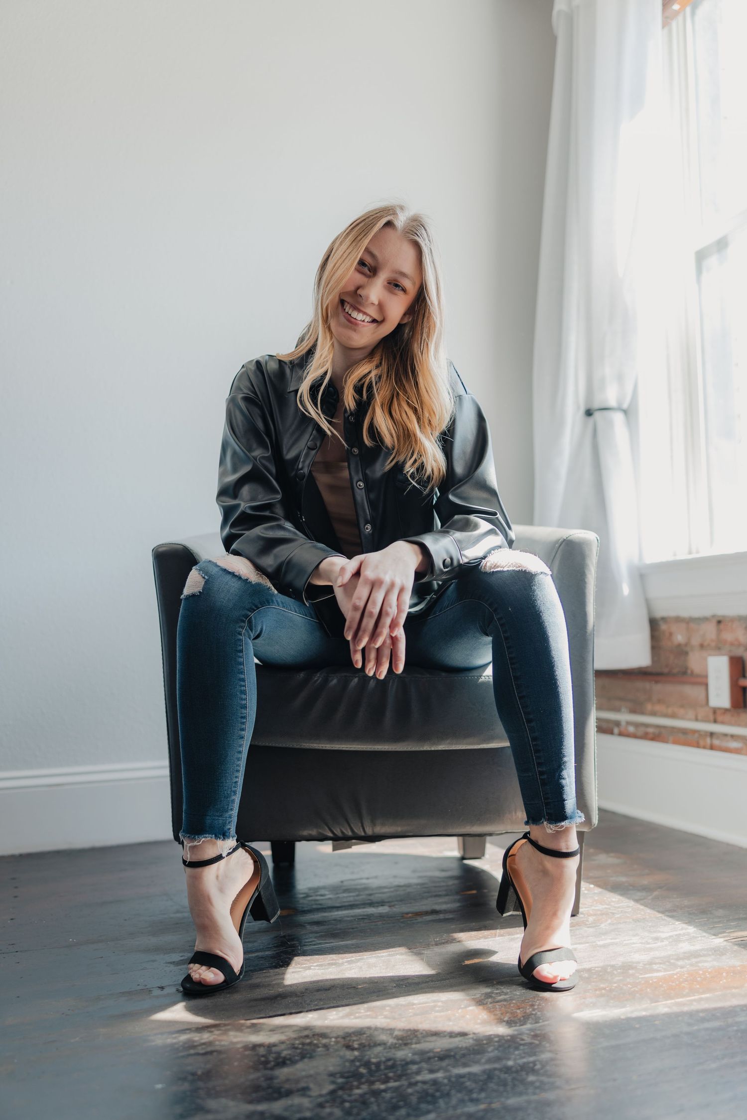 Smiling blonde woman in black jacket and jeans sitting in a chair by a bright window.