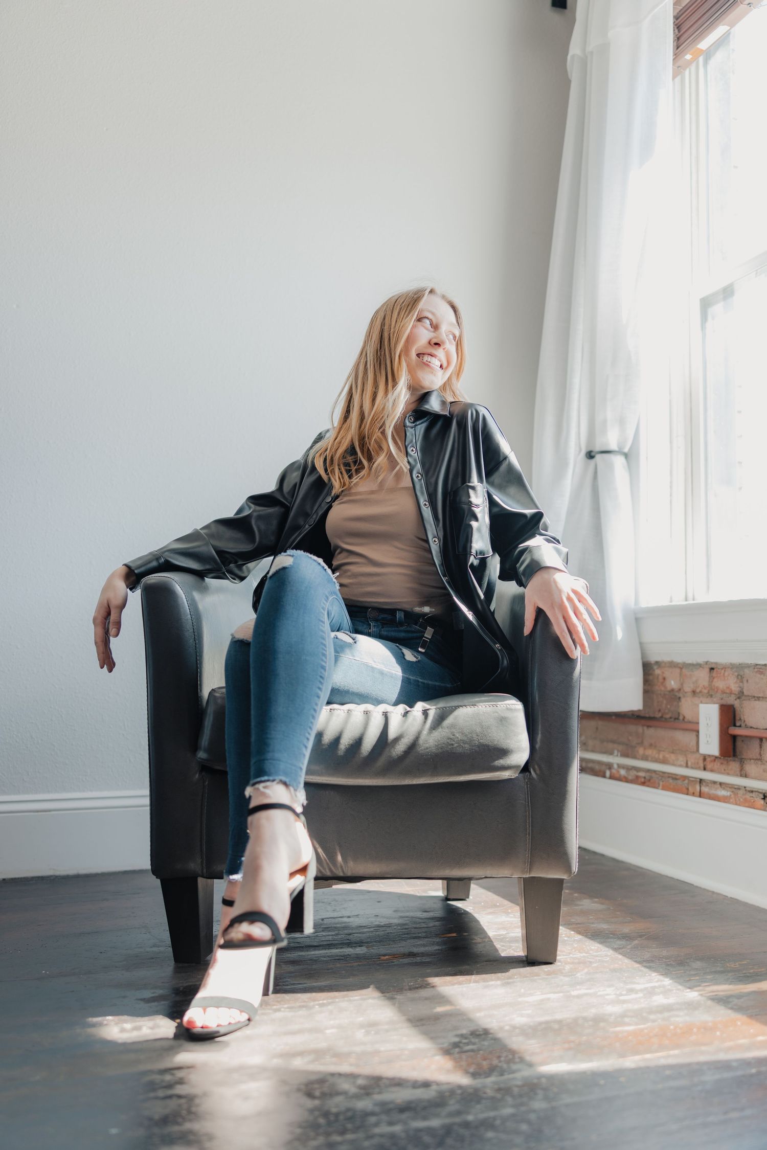 Smiling blonde woman in ripped jeans and black jacket sitting relaxed in a dark leather armchair by a sunlit window.