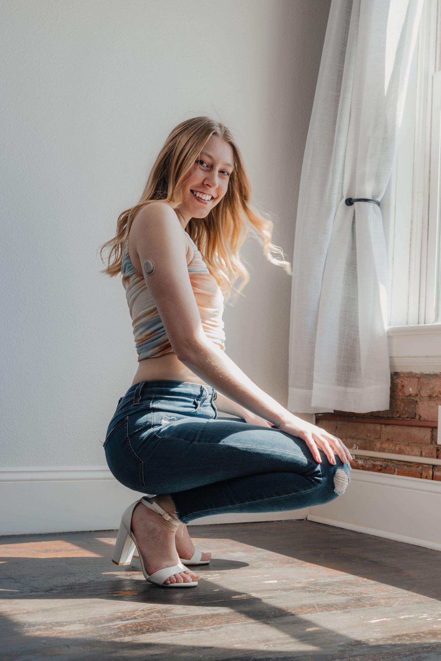 Smiling young woman with blonde hair crouching in blue jeans, crop top, and white heels near a sunlit window.