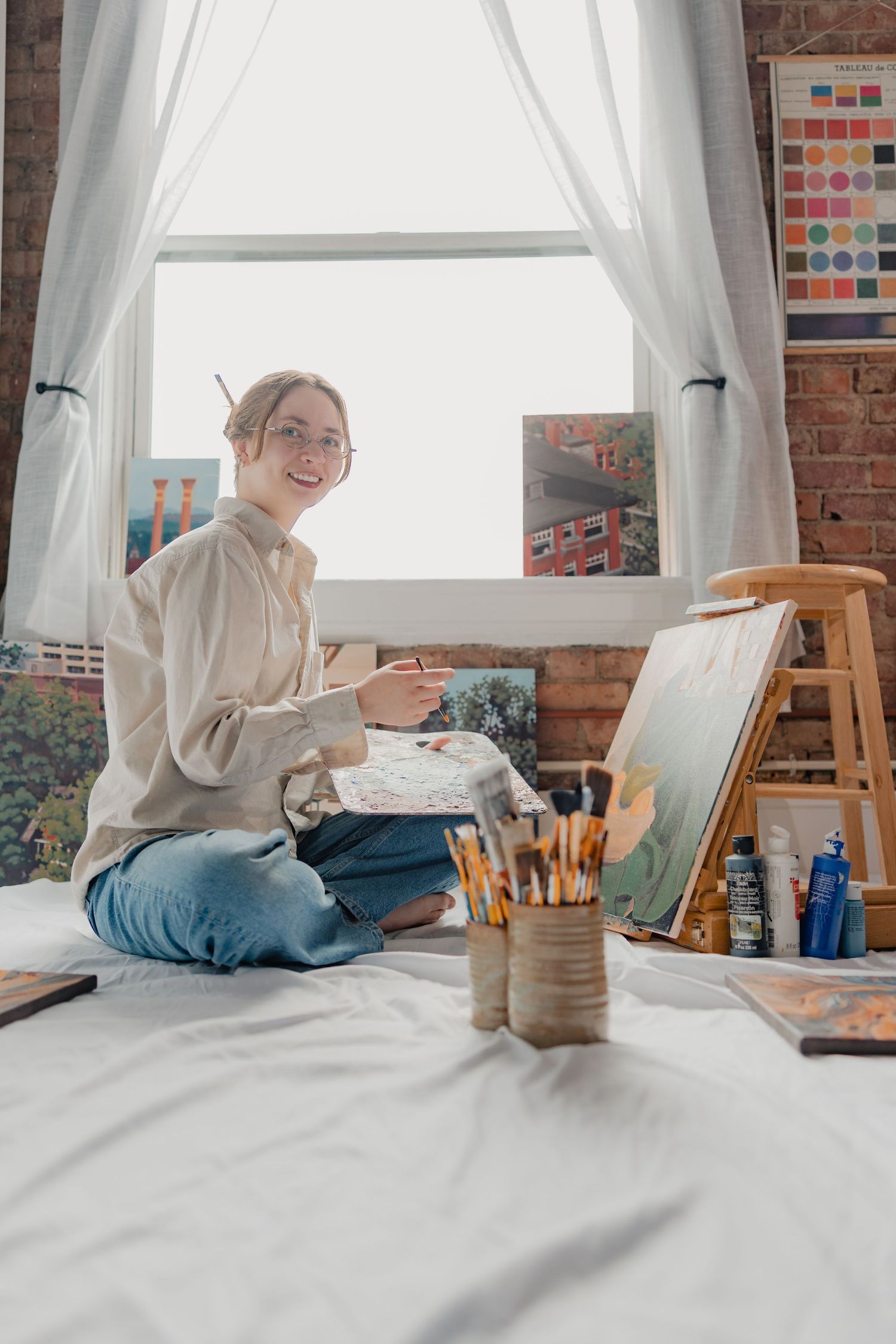 Young female artist sits cross-legged on white sheet, painting on easel near brick wall window in cozy art studio.