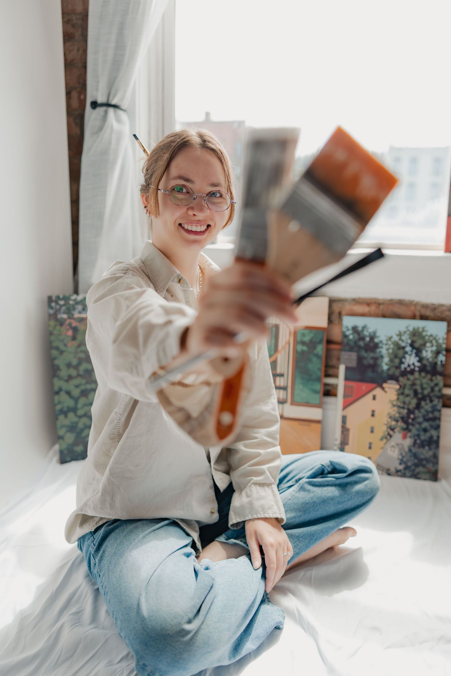 Smiling female artist sitting cross-legged by window, holding paintings, wearing glasses and casual linen shirt in bright studio.