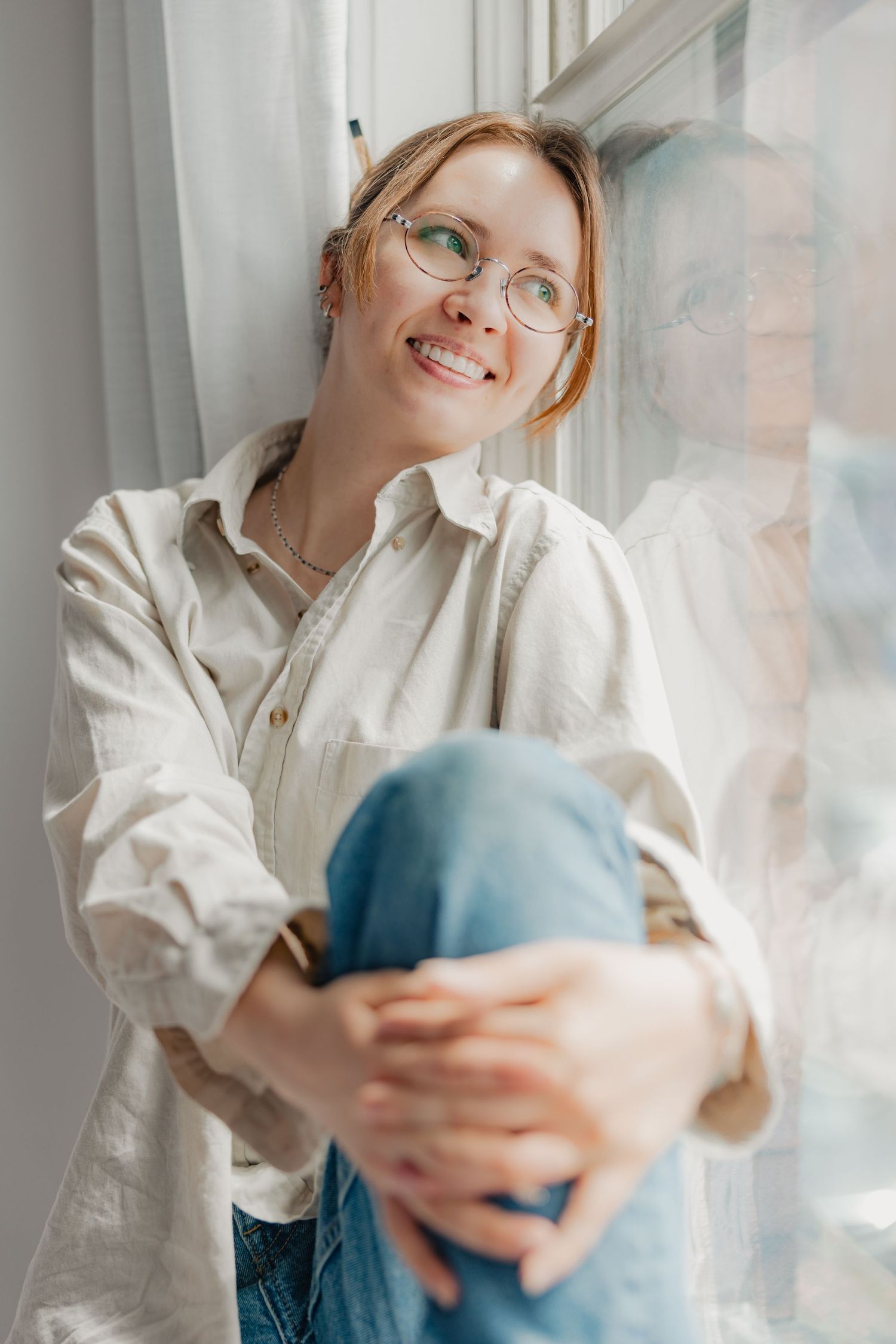 Smiling young woman with glasses and pencil in hair sits by window, hugging knees, wearing white shirt and jeans.