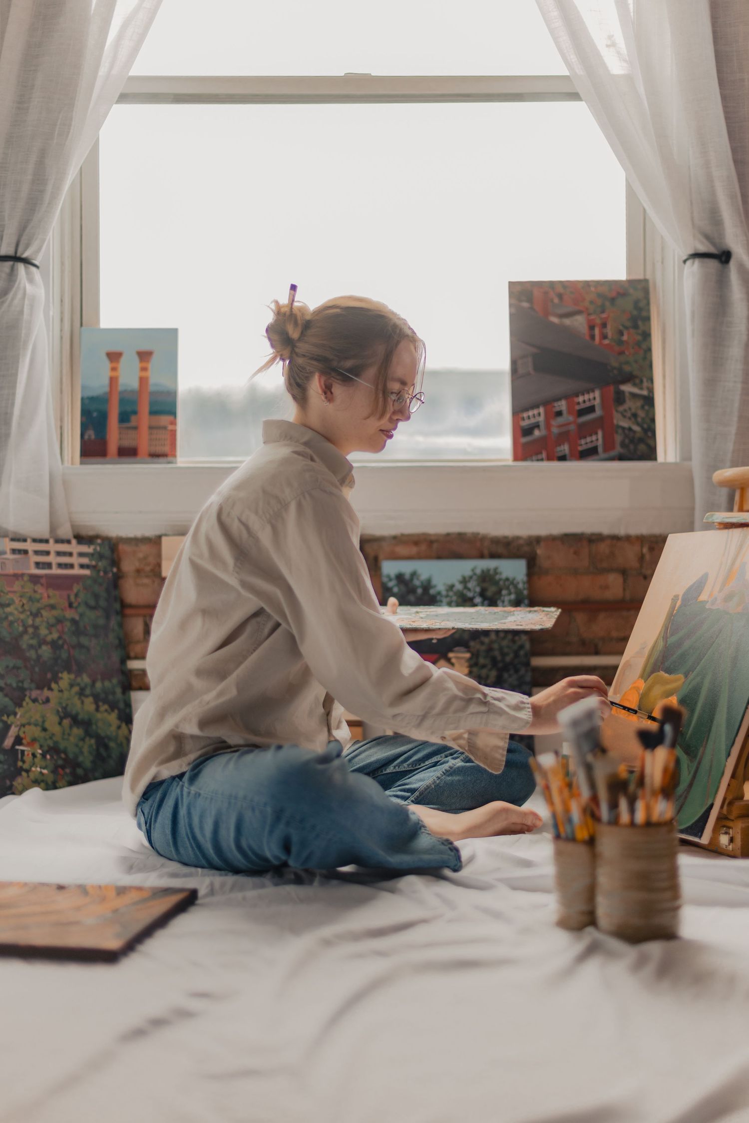 Young woman artist sitting cross-legged on bed painting on canvas near bright window with artwork displayed behind her.
