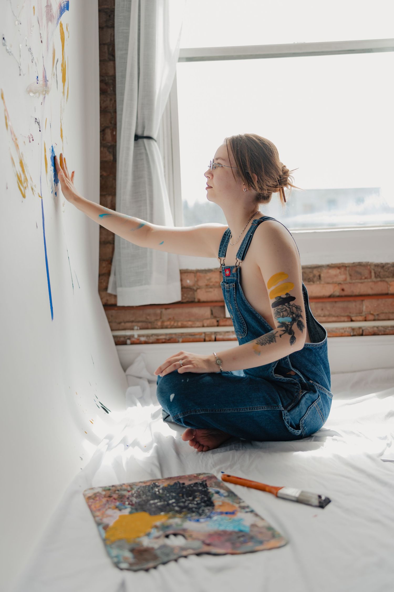 Young female artist in denim overalls painting on a white canvas wall, sitting cross-legged with a colorful palette nearby.