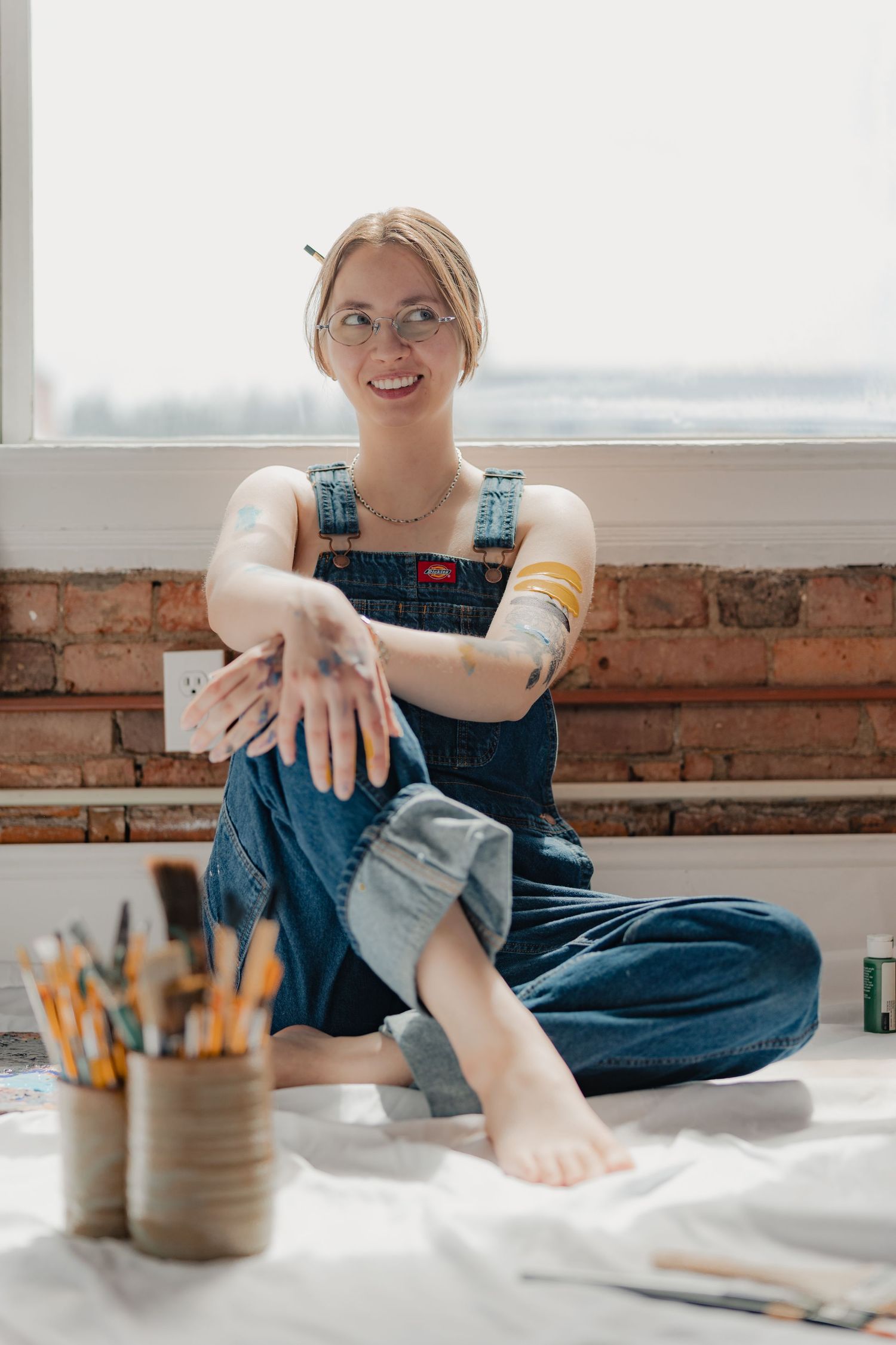 Smiling female artist in denim overalls sits cross-legged on floor with paint brushes nearby in a bright studio.