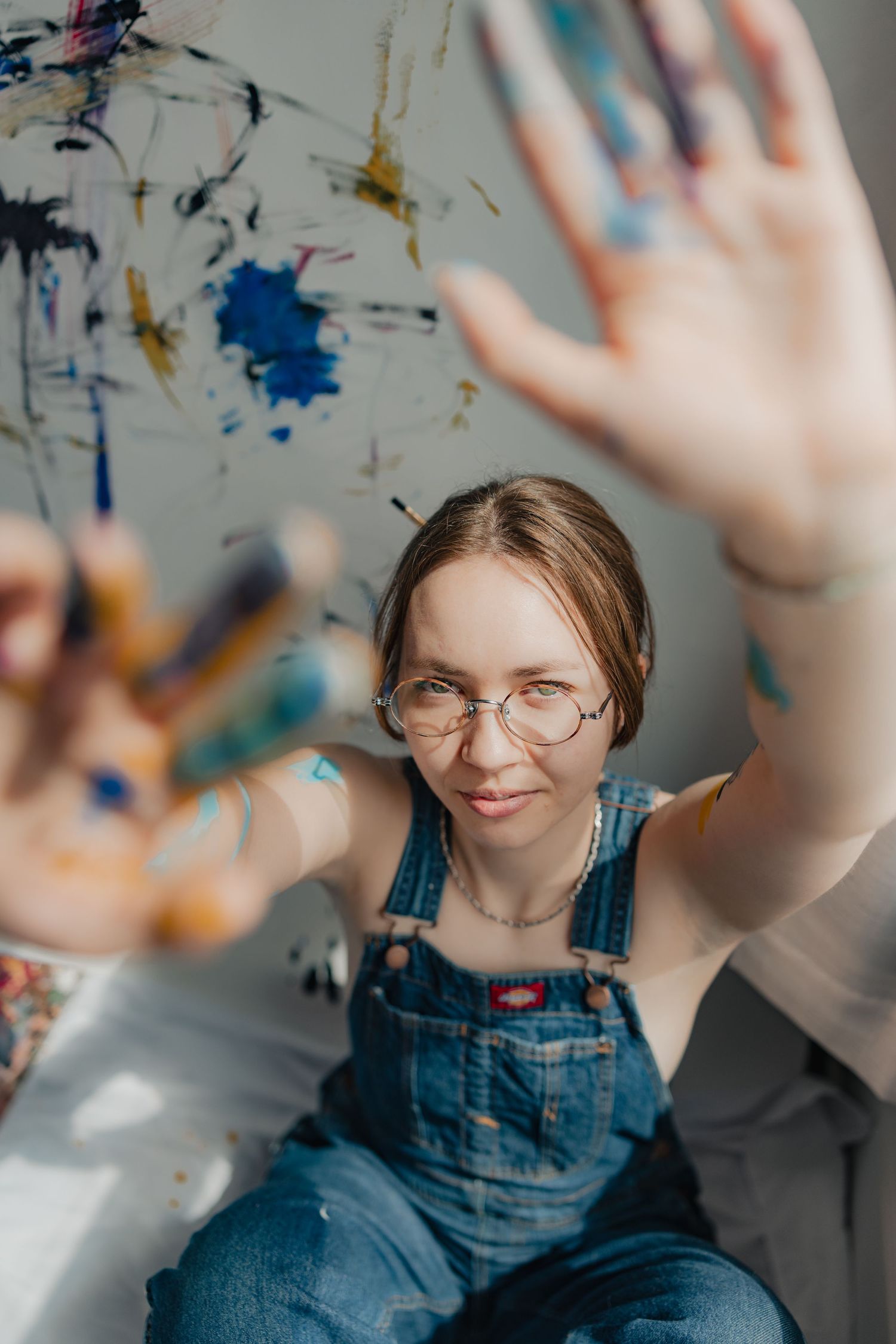 Female artist in denim overalls shows paint-covered hands toward camera in front of colorful abstract artwork.