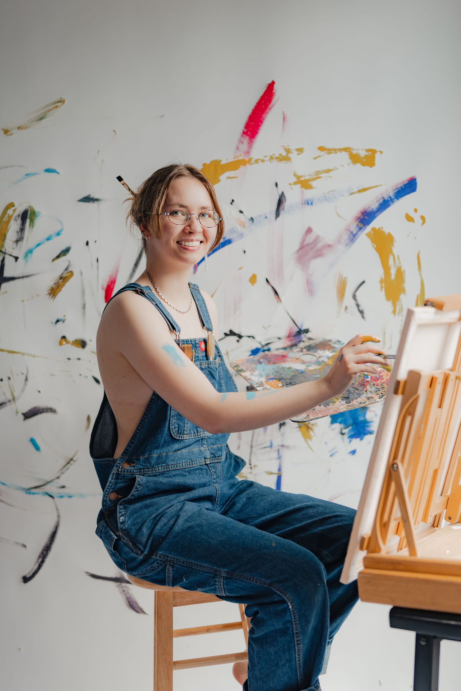 Smiling female artist in denim overalls painting on easel in art studio with colorful abstract paint splashes on wall.