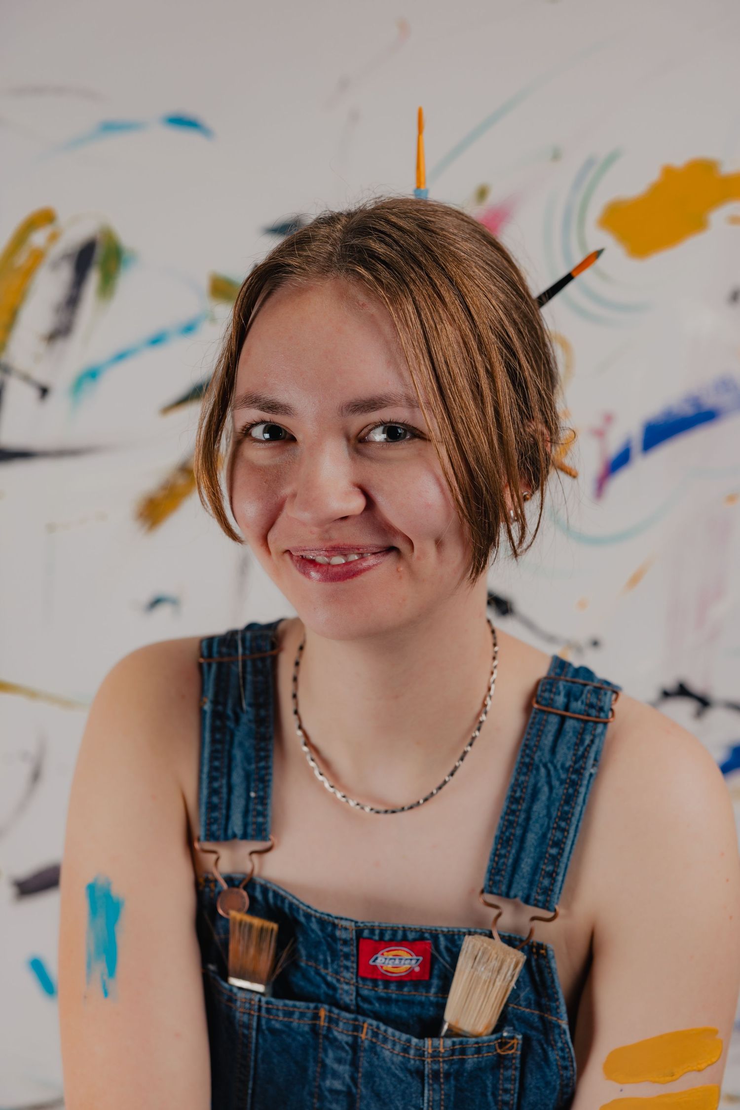 Smiling young female artist in denim overalls with paintbrushes in hair, paint-covered hands, in front of colorful artwork.