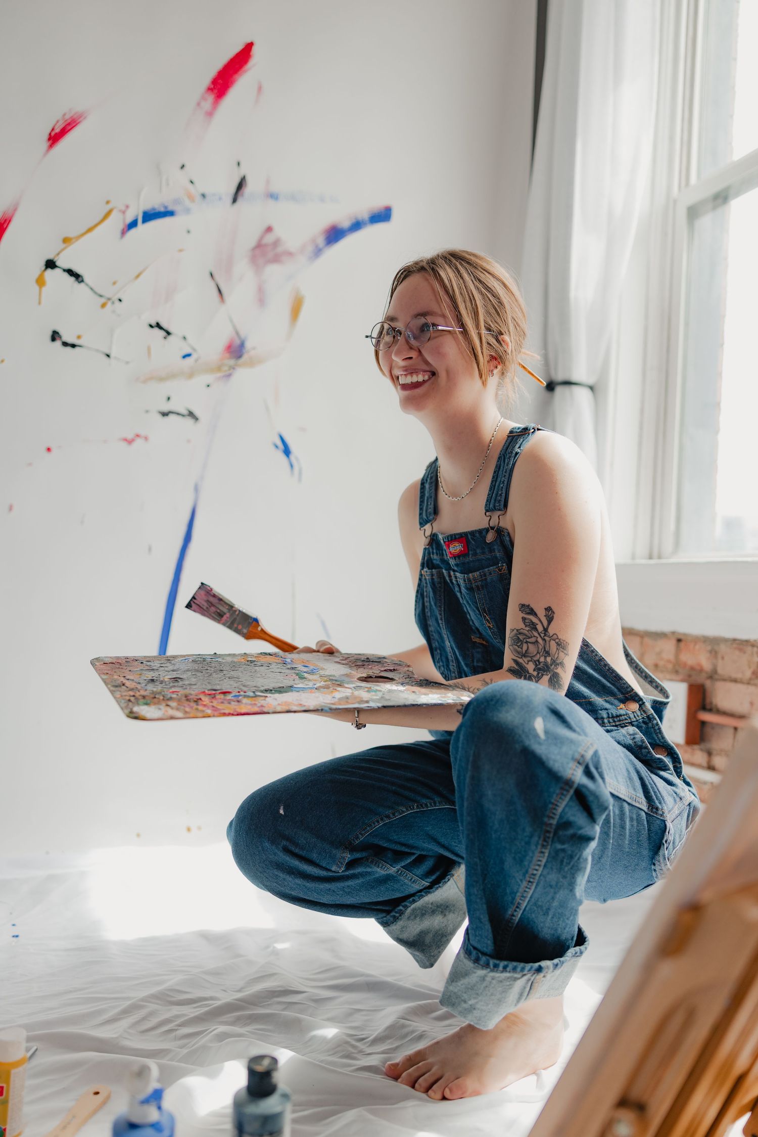 Smiling female artist in denim overalls holds paint palette in a colorful splattered studio.