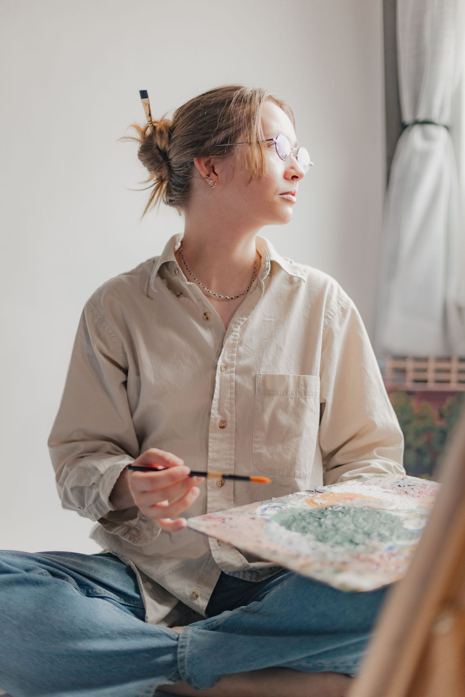 Young female artist sitting cross-legged, holding a paintbrush and palette, wearing glasses and a beige shirt.