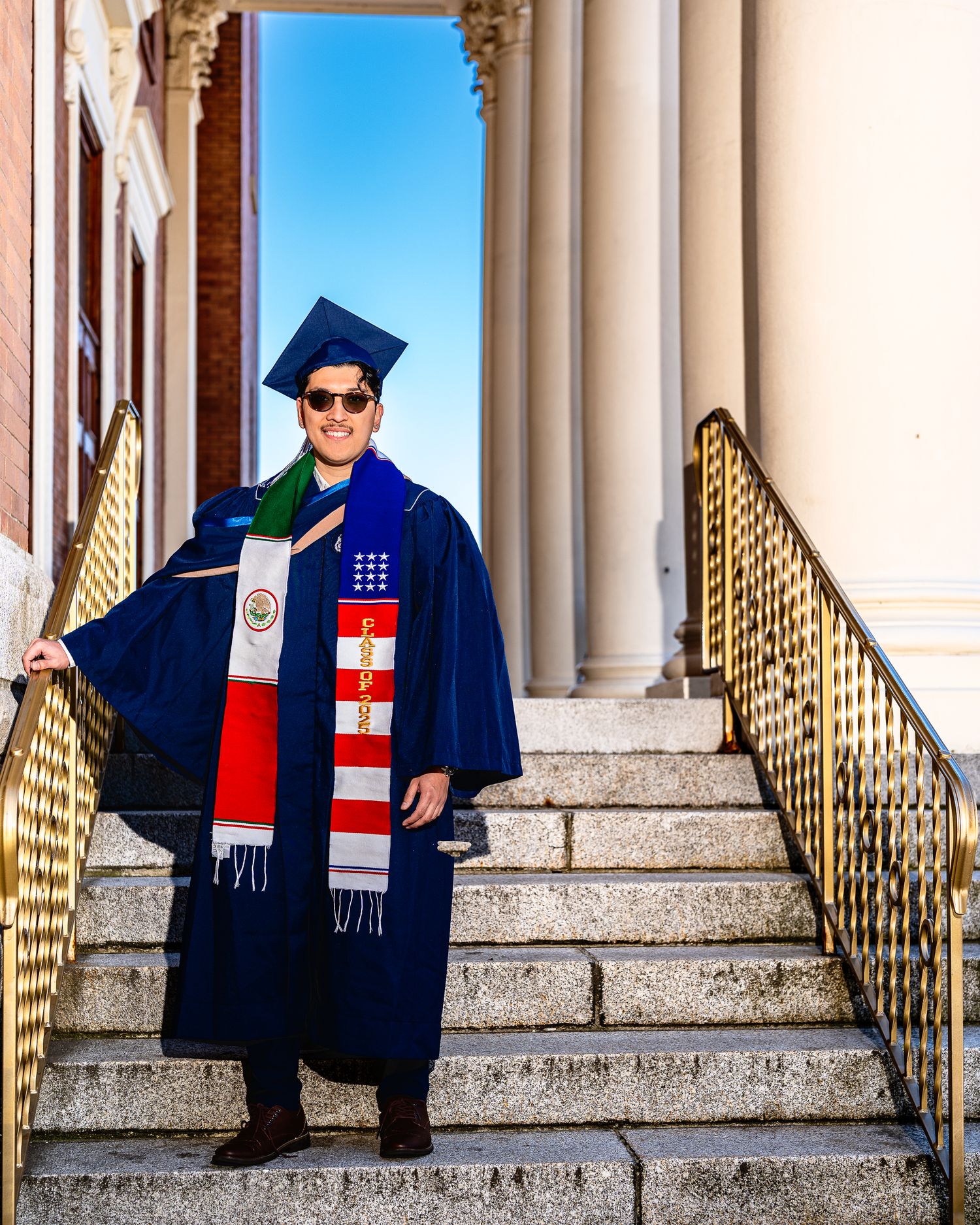 Graduate in blue academic regalia with national flag scarves sits on marble steps of ornate building.