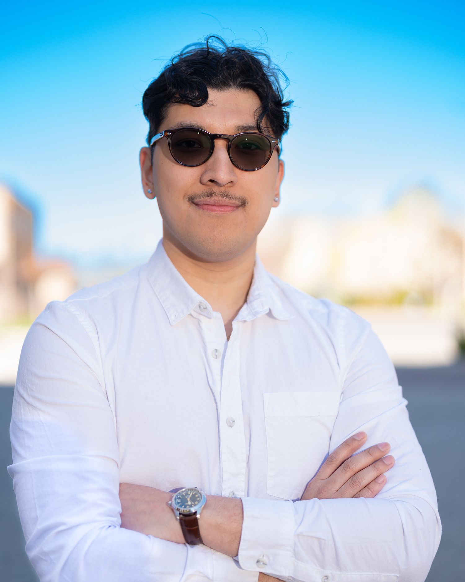 A person in a white button-down shirt and sunglasses poses confidently against a bright blue sky background.