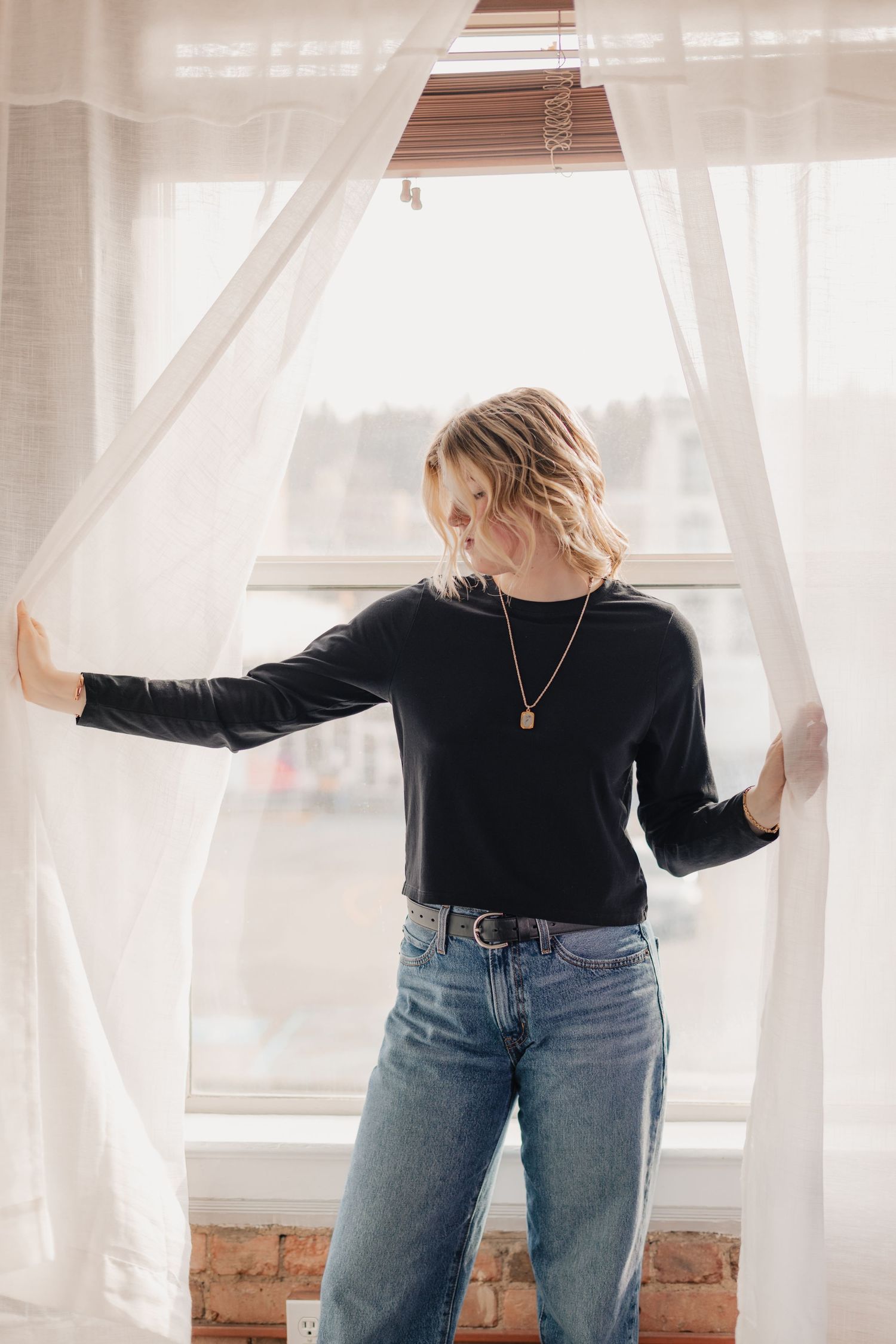 A person in a black sweater and jeans stands by a window with flowing white curtains in natural light.