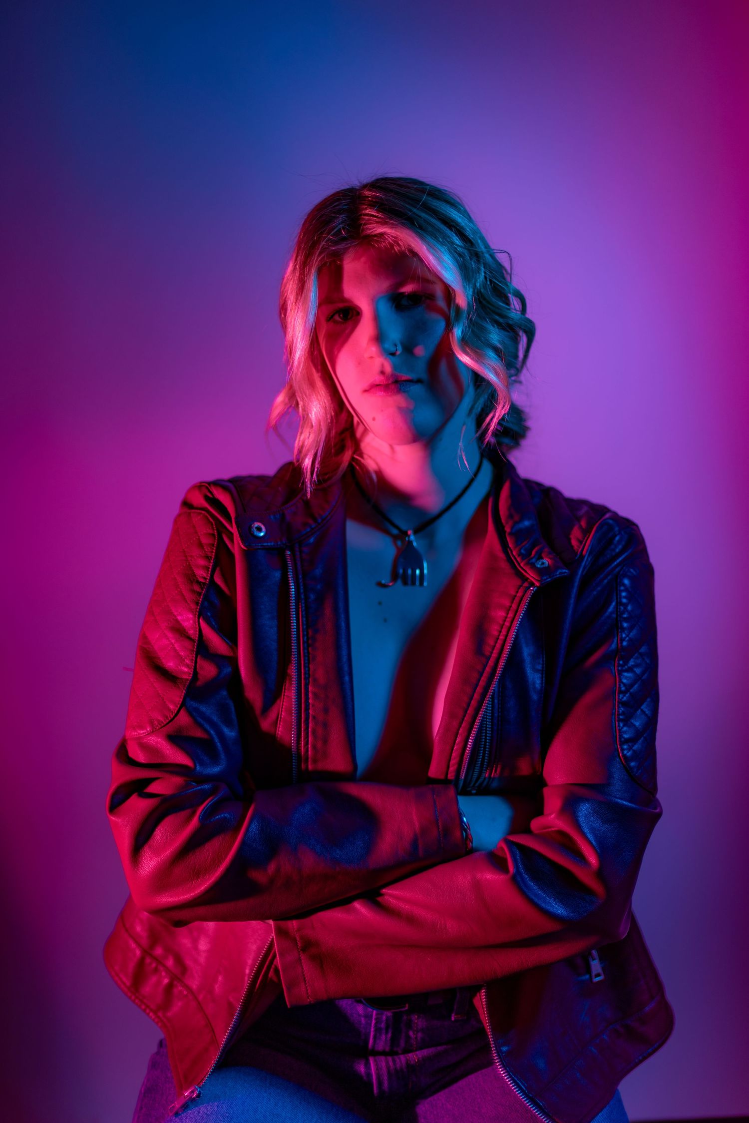 Moody portrait with dramatic red and blue lighting against purple background showing figure in leather jacket with crossed arms.