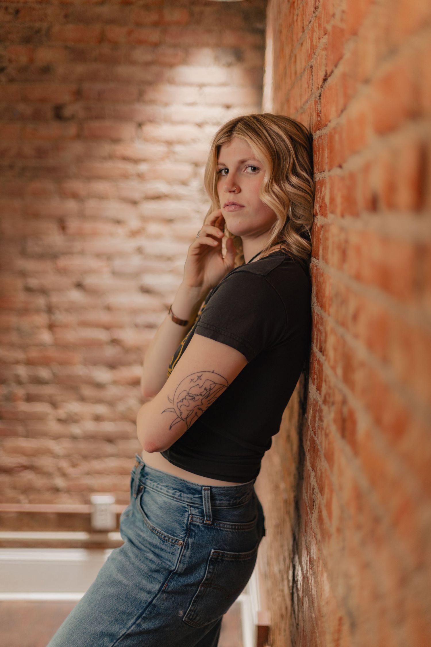 Person in black top and jeans poses against rustic brick wall in artistic portrait series.