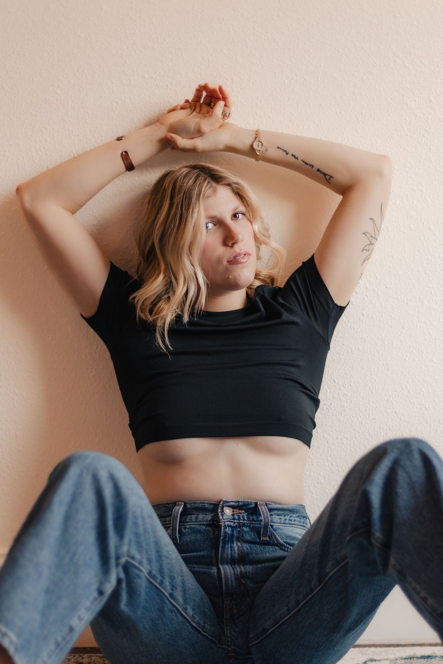 A person in a black crop top and blue jeans poses casually against a beige wall with arms raised overhead.