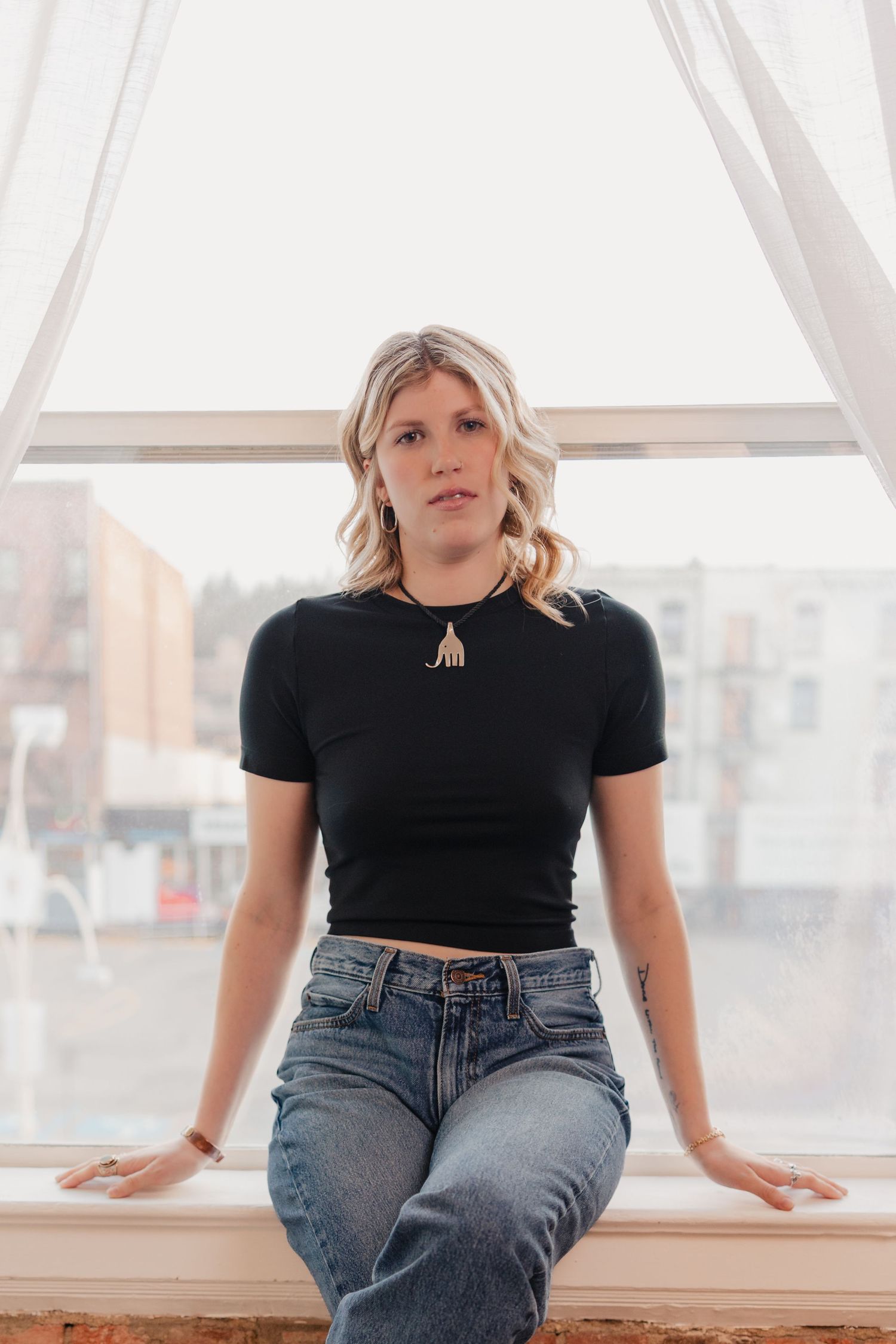 Person in black crop top and blue jeans sitting casually on windowsill with city view in background.