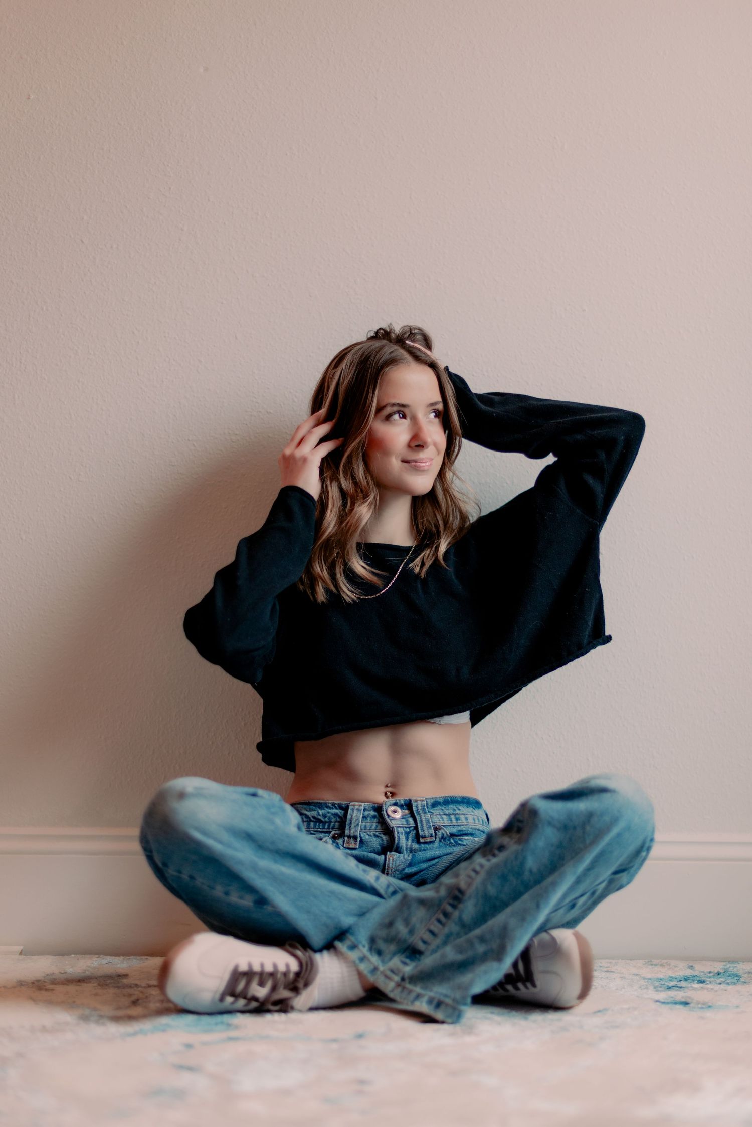 Candid portrait sitting cross-legged on floor wearing black sweater and jeans with wavy hair.