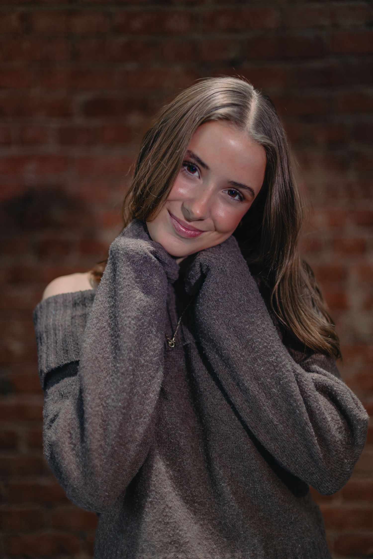 Cozy portrait against brick wall wearing oversized grey sweater with hands near face.