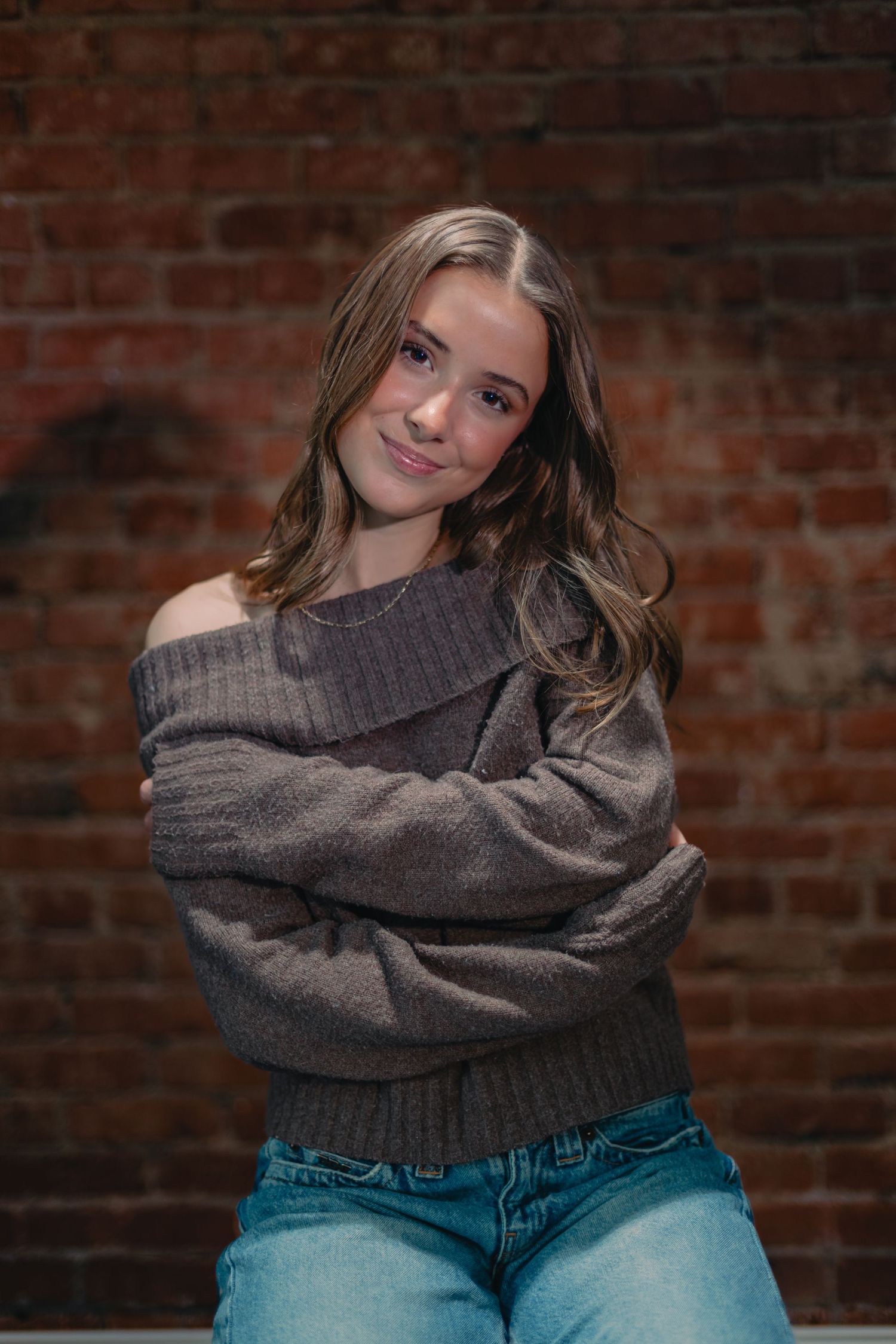 Casual portrait wearing grey knit sweater and jeans against textured brick wall.