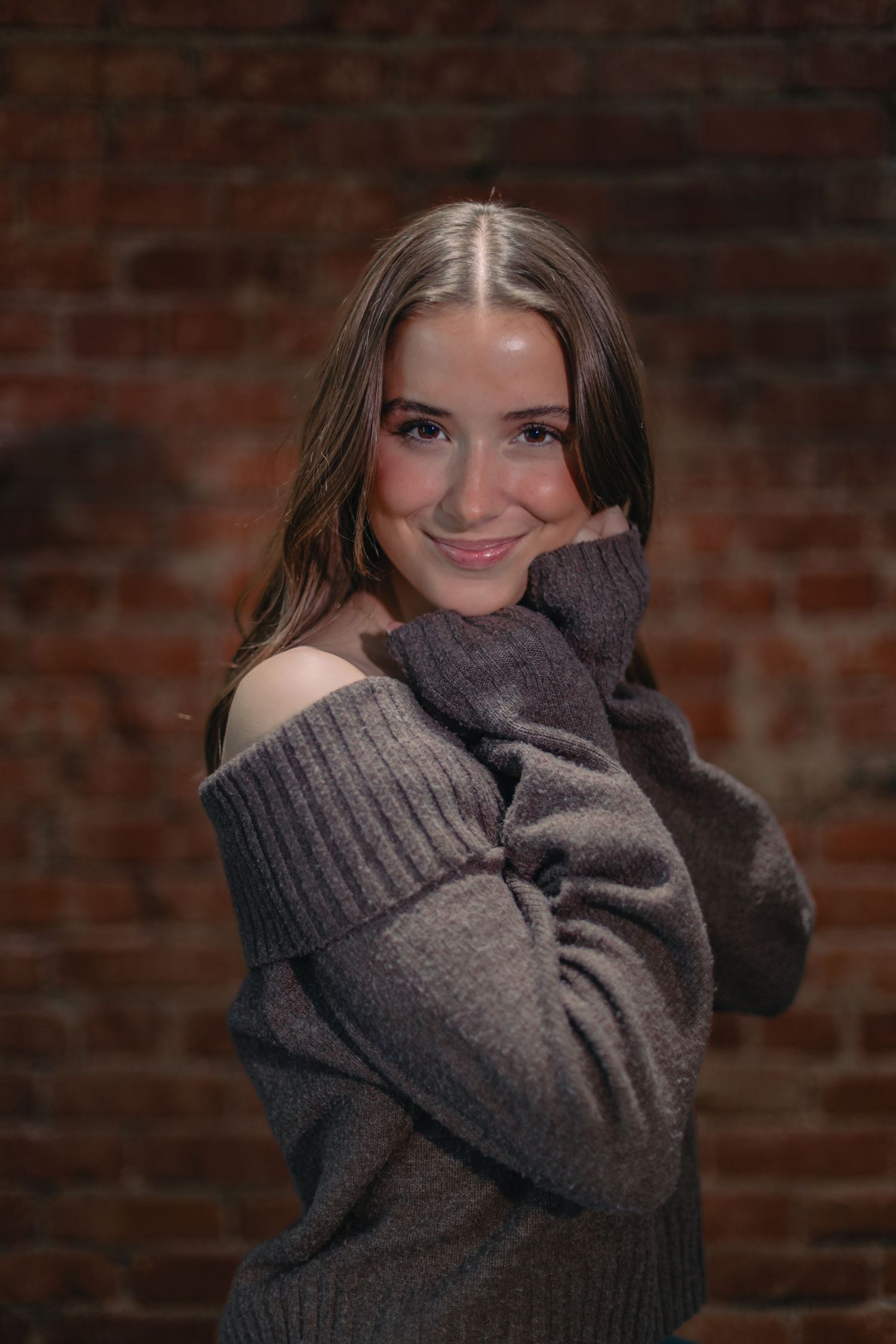 Lifestyle portrait in grey off-shoulder sweater against rustic brick background.