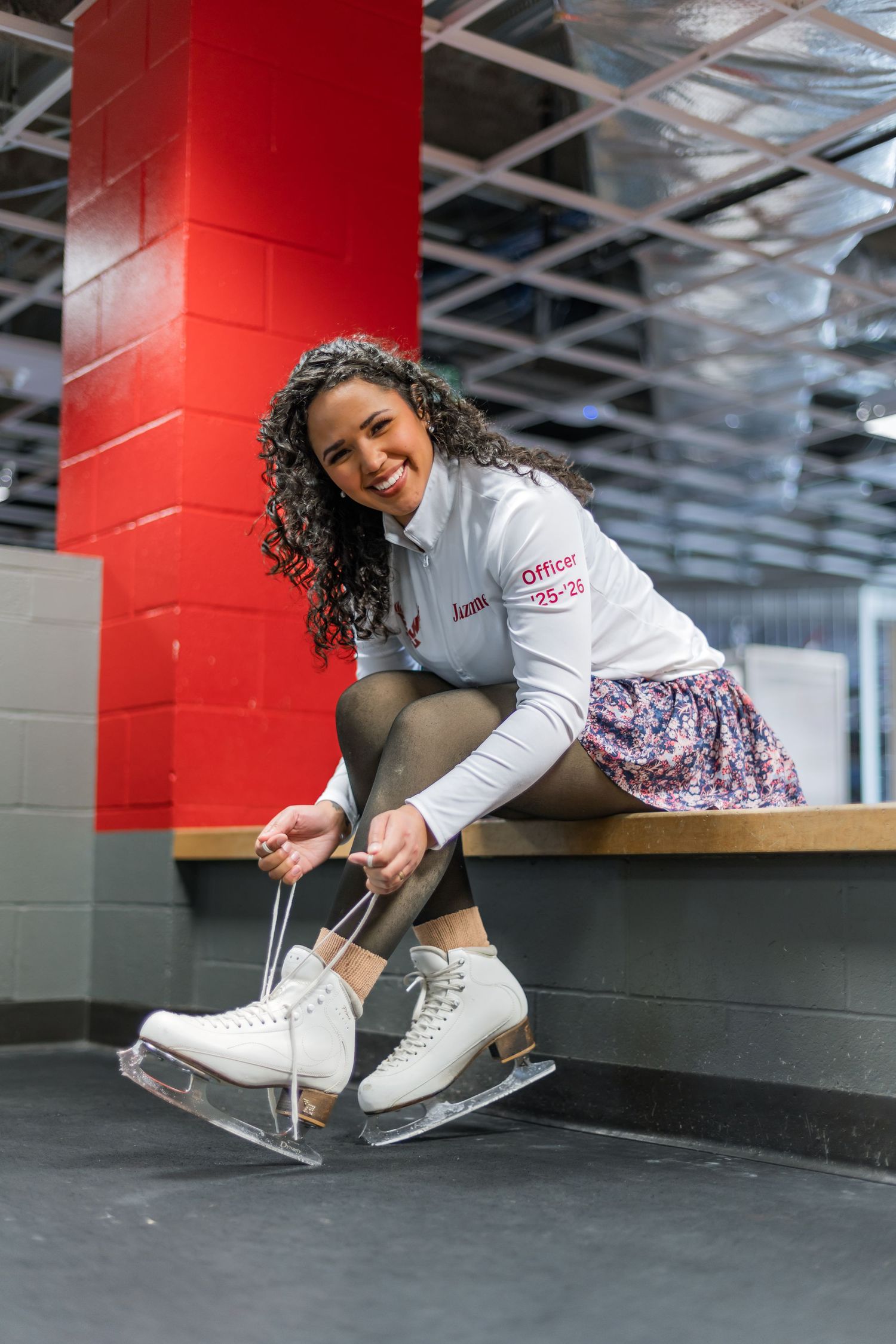Smiling woman with curly hair lacing white figure skates on a bench at an indoor ice skating rink.