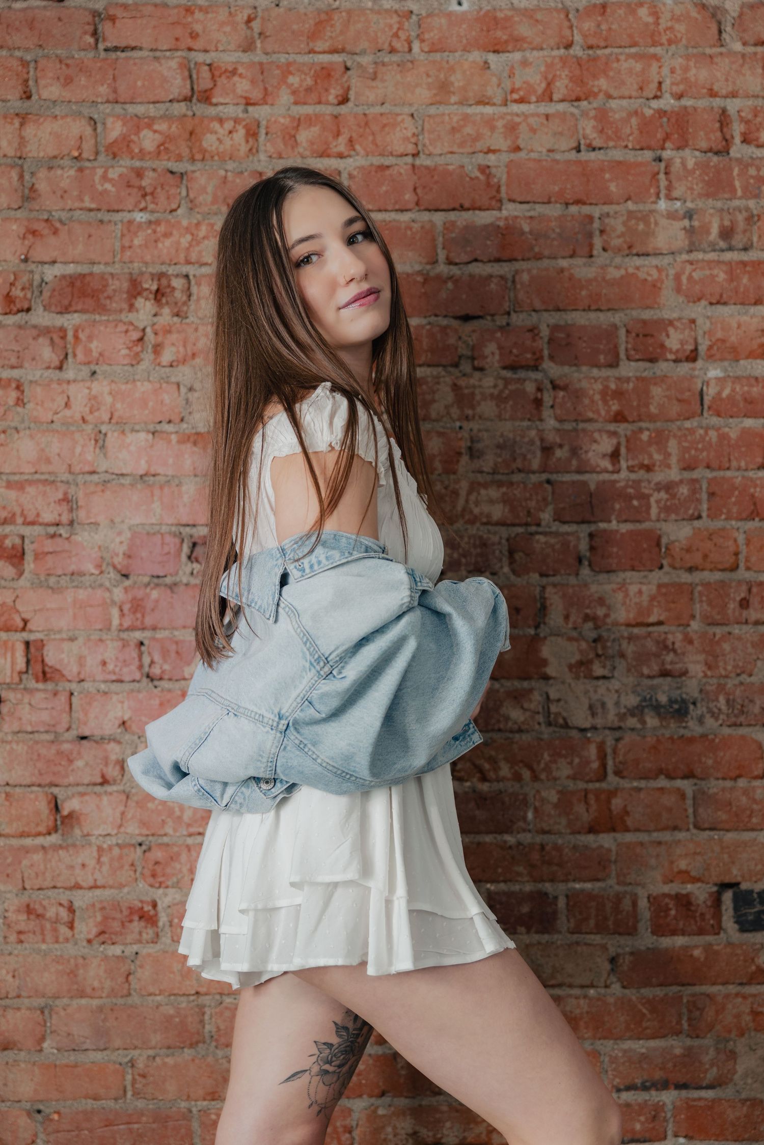 Fashion model poses against rustic brick wall wearing light denim jacket and white dress in artistic photoshoot series.