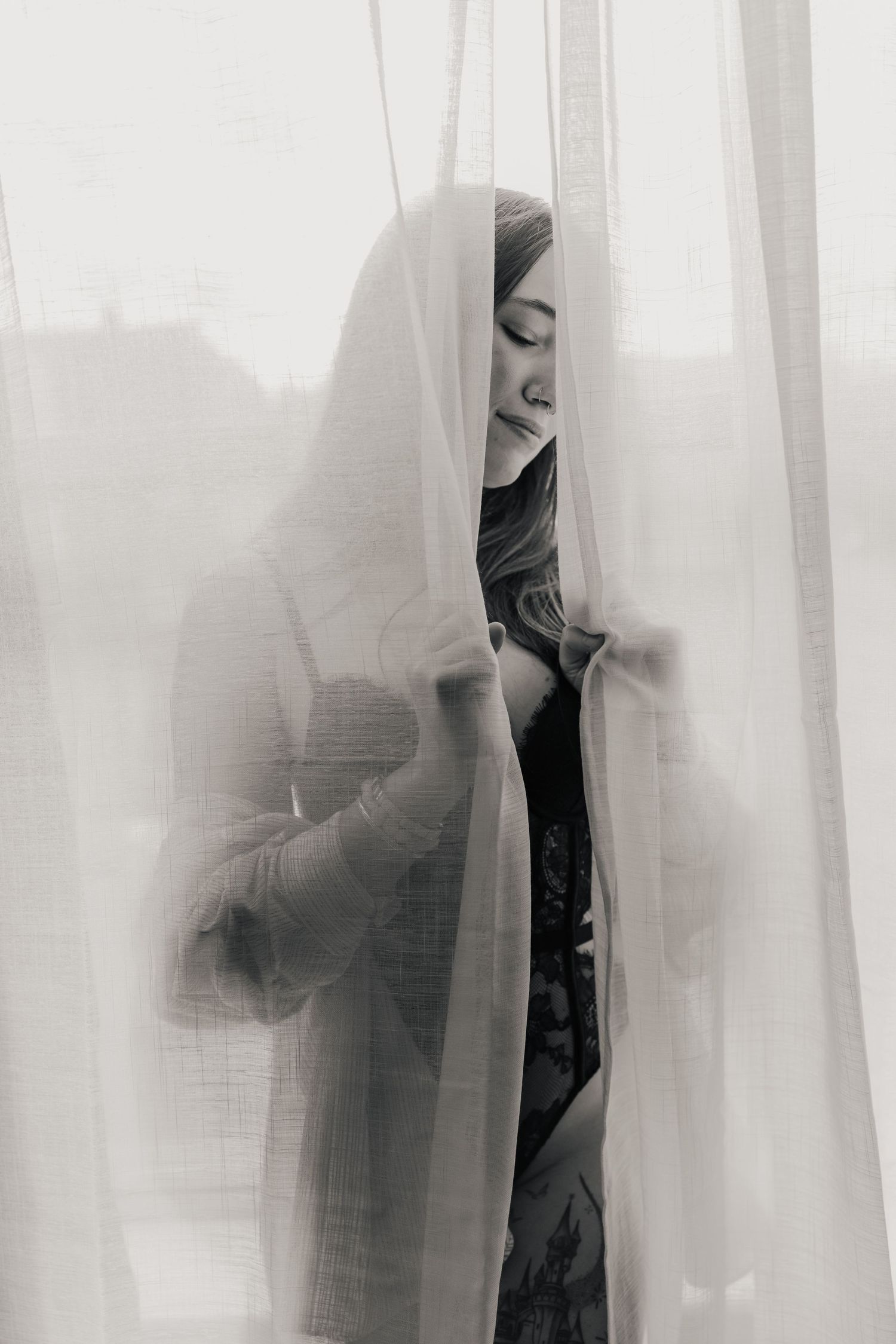 Silhouetted figure visible through sheer white curtains in a moody black and white artistic composition.