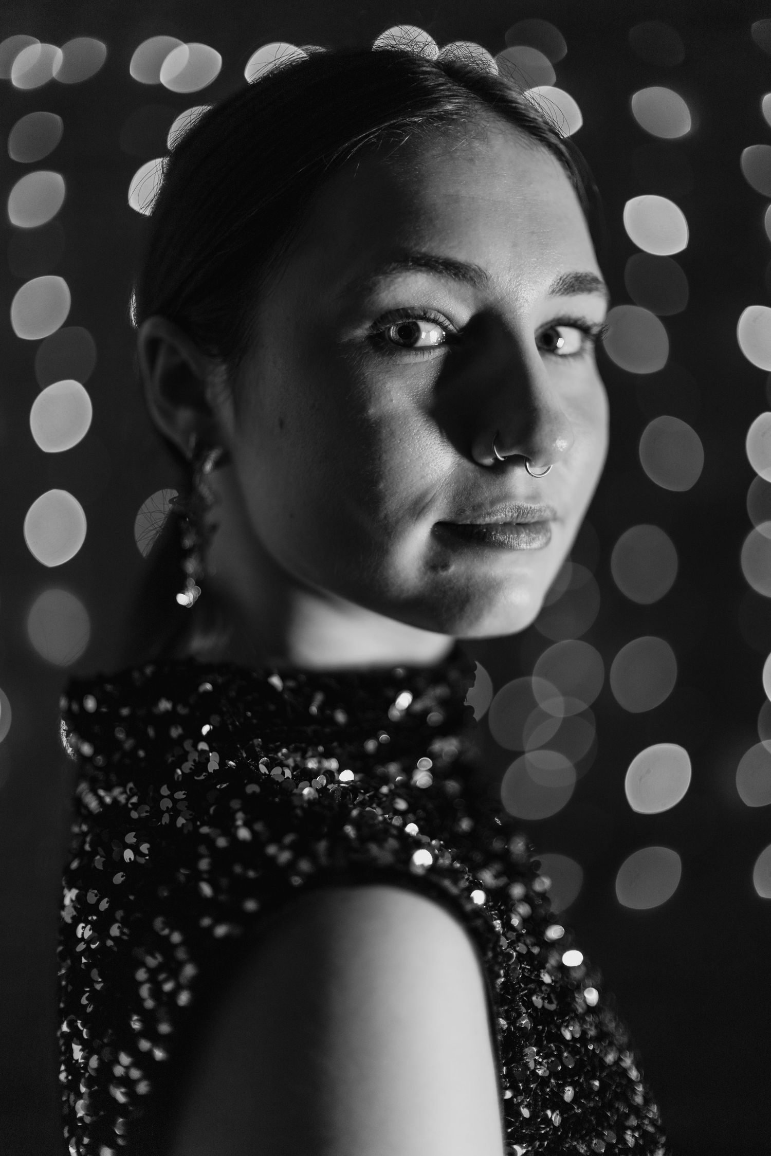 Black and white portrait with bokeh lights showing a person wearing a sequined dress and nose piercing against dark background.