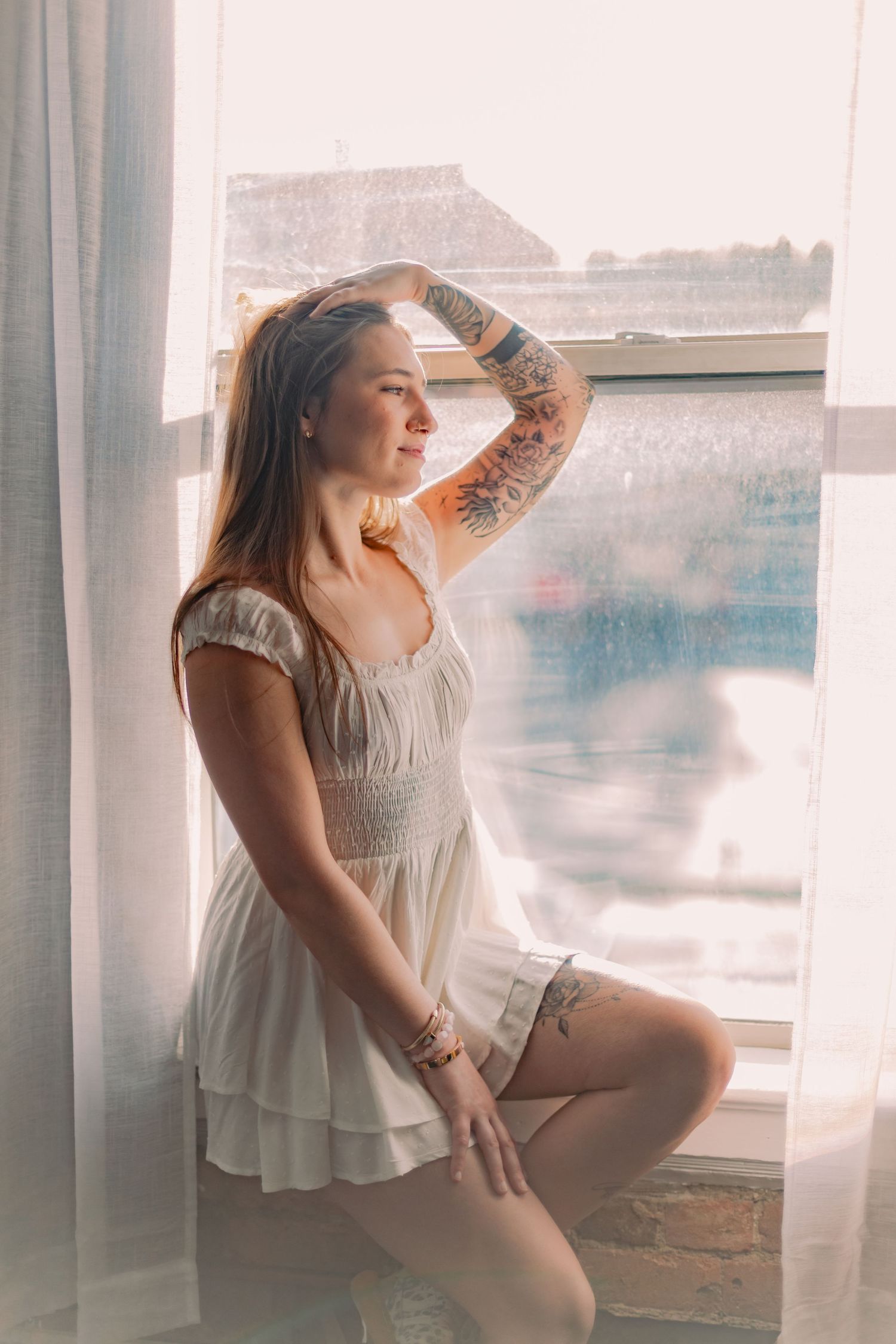Dreamy boudoir photo series of someone in a white dress posing by a sunlit window.