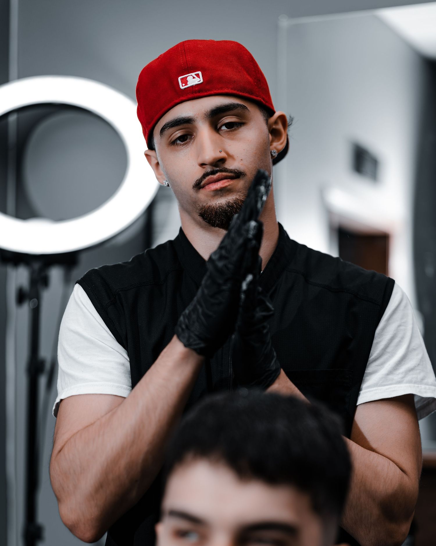 A person wearing a red MLB cap, white t-shirt and black vest poses while wearing black gloves in a salon setting.