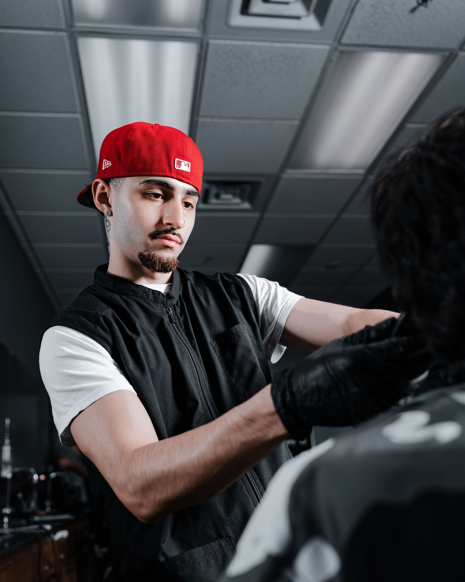 A barber wearing a red cap and black vest works diligently in a dimly lit salon with fluorescent lighting overhead.