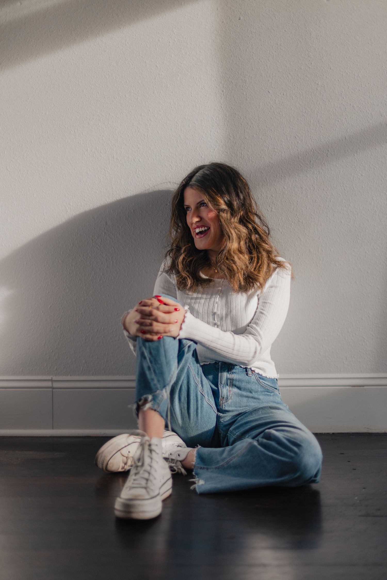Person sitting on dark floor wearing white sweater and baggy jeans with white sneakers, laughing joyfully in natural light.