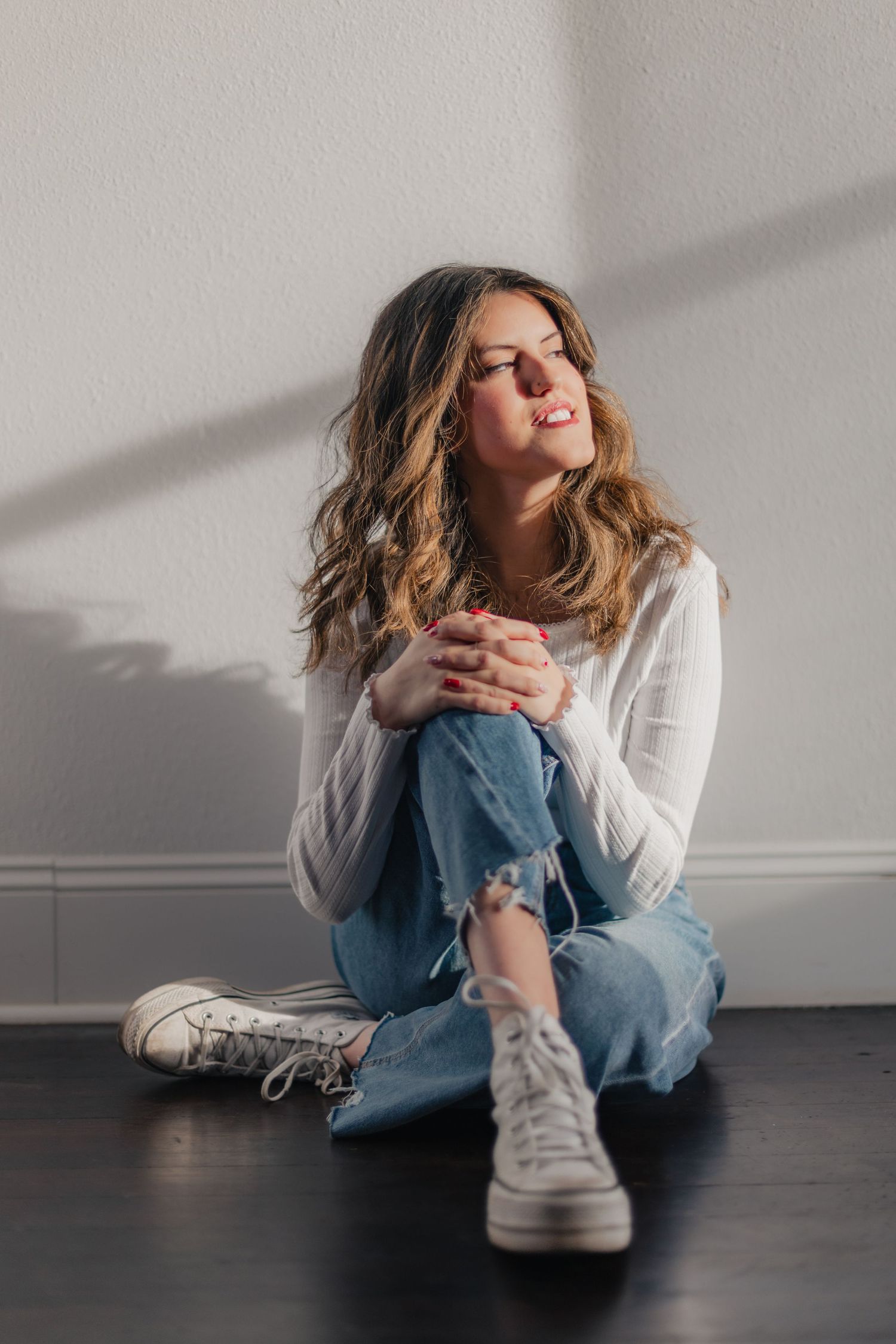 Person in white sweater and jeans sits on dark floor against wall with dramatic shadows and lighting.