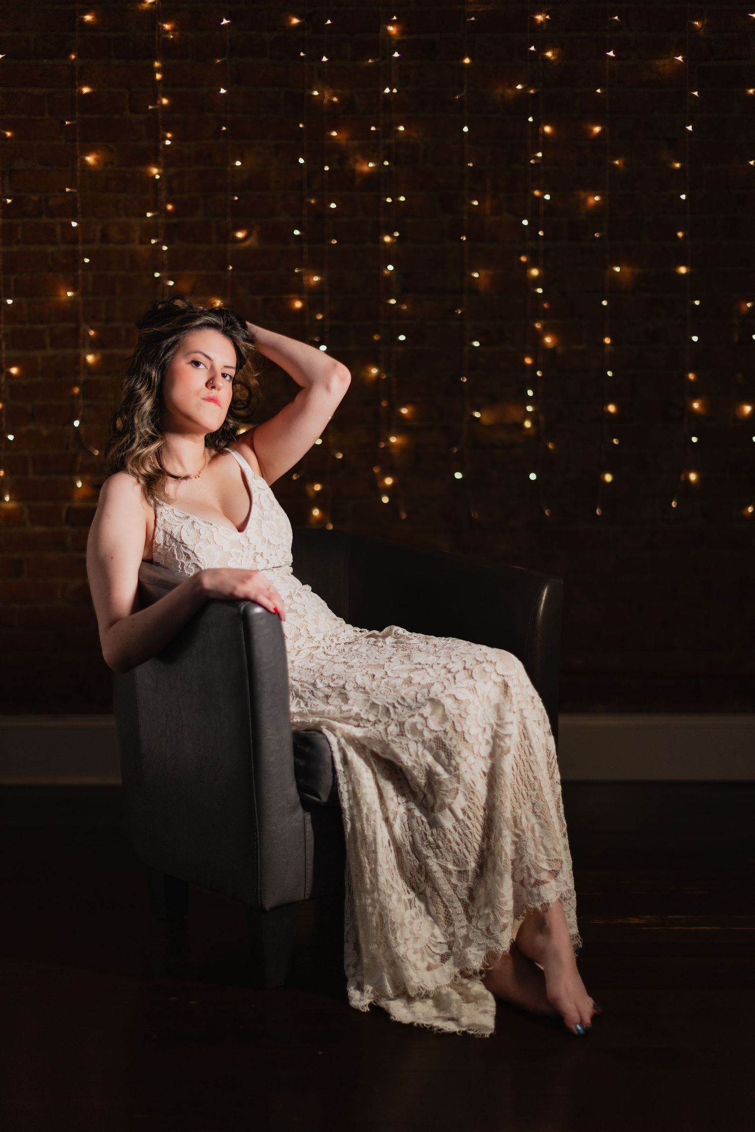A person in a lace wedding dress poses on a chair against a backdrop of twinkling string lights.