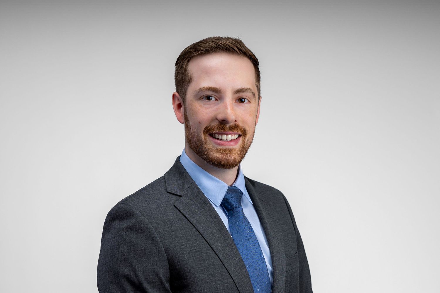 Professional headshot of someone wearing a gray suit and blue tie against a light gray background.