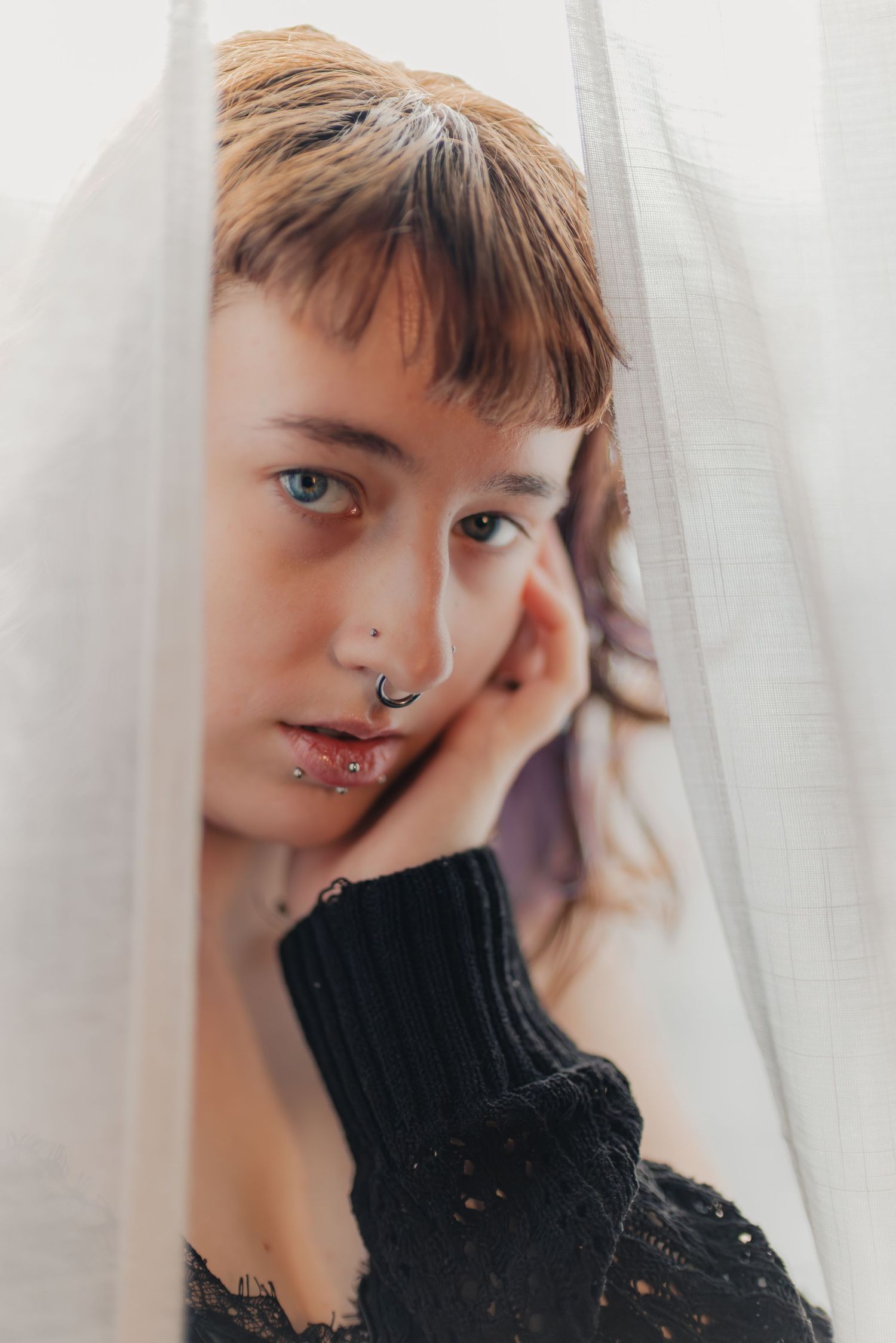 Close-up portrait showing multiple facial piercings and a black turtleneck sweater against a white curtained background.