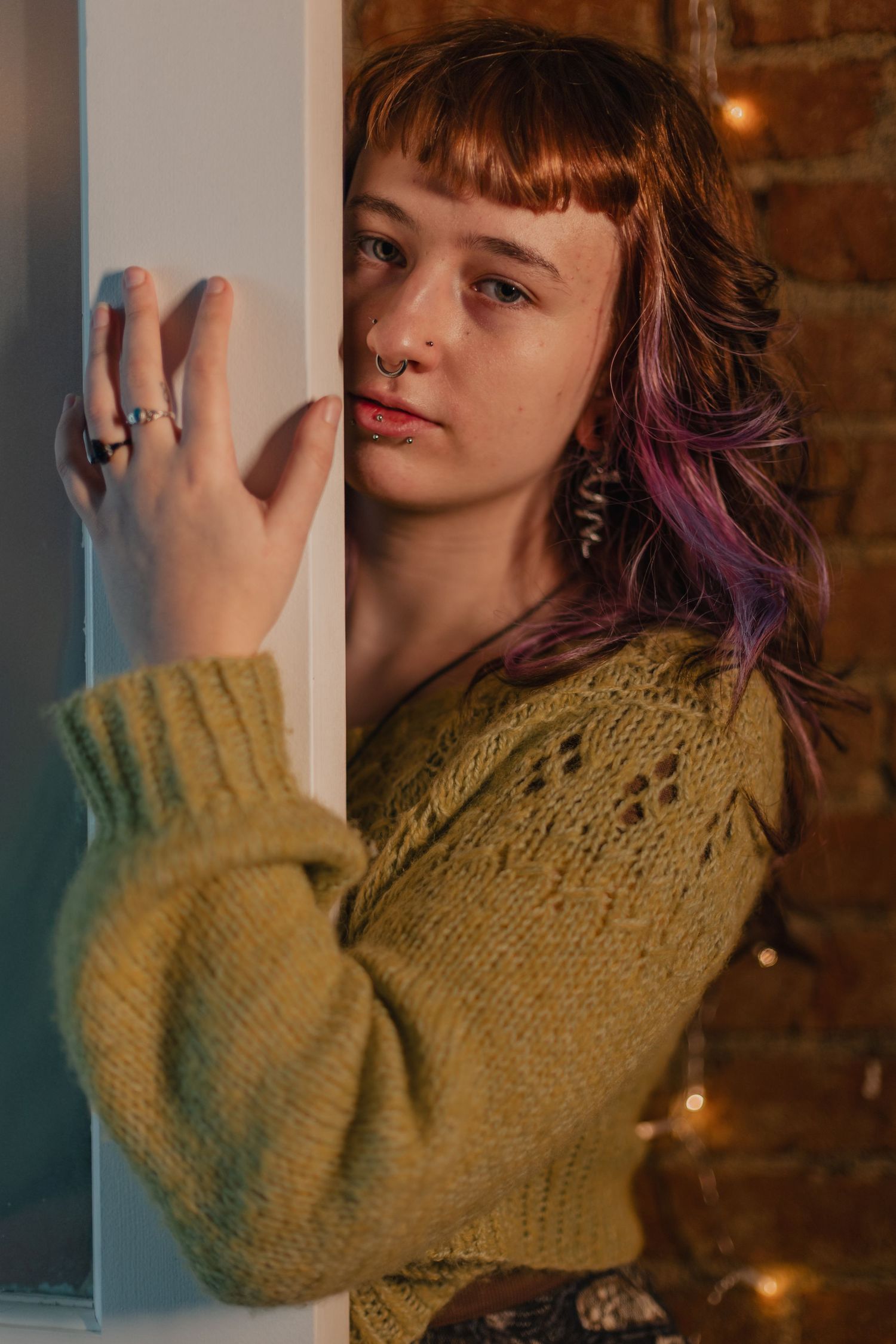 A person with colorful hair wearing a yellow knit sweater poses against a brick wall with soft lighting in the background.