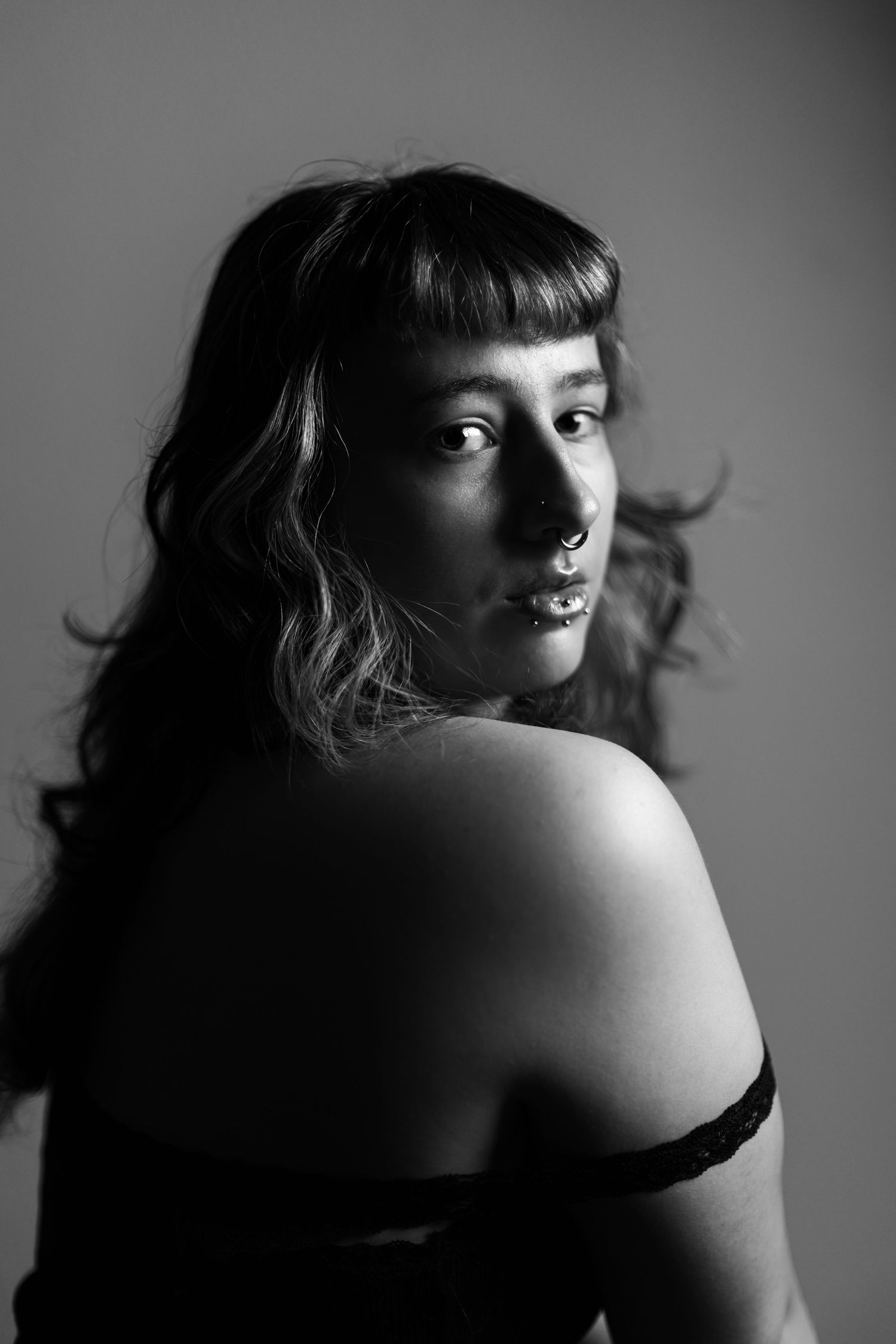 Dramatic black and white portrait of a person with bangs looking over bare shoulder in moody lighting.