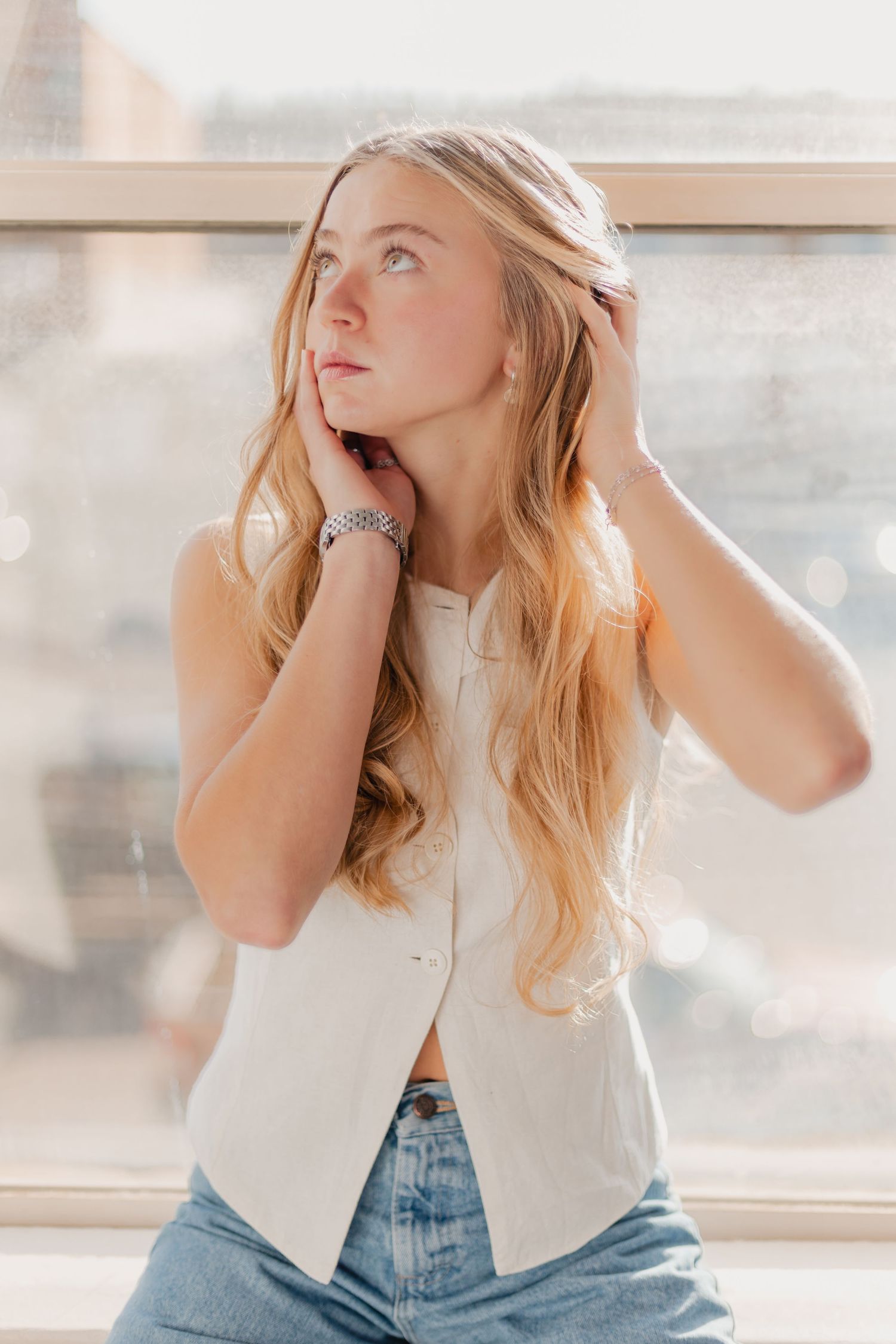 A person with blonde hair poses by a window in a white top and jeans while making expressive gestures.